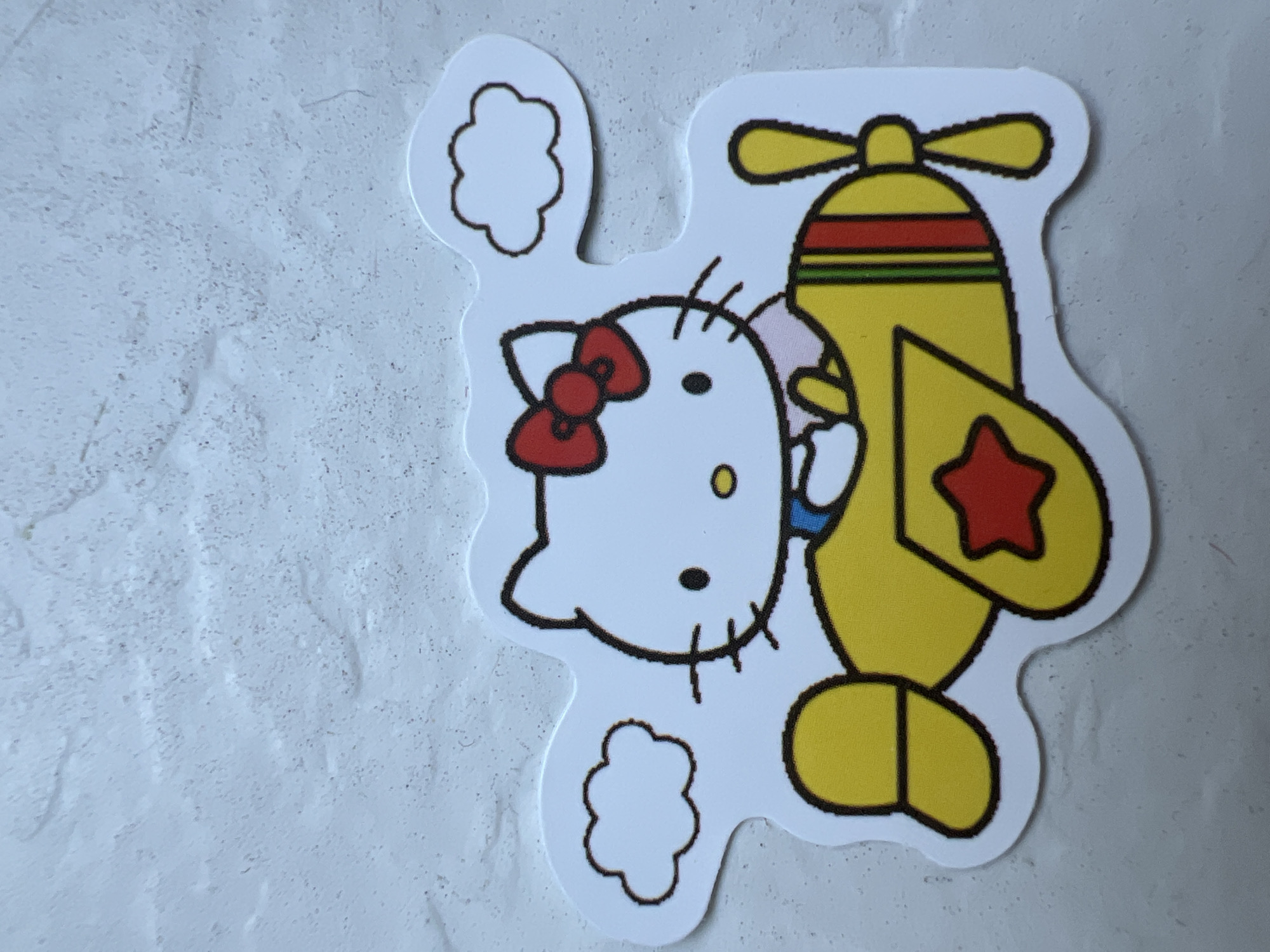 A sticker of Hello Kitty riding in a yellow airplane is placed on a textured white surface. The sticker depicts Hello Kitty wearing a blue shirt and a pink collar, with her signature red bow on her head. The airplane has a red star on its side and multi-colored stripes. Two small, simple cloud shapes are attached to the sticker, one to the left and one to the right of Hello Kitty's head. The background is a light gray, textured surface resembling a wall. There is no indication of any specific location in Warsaw, Poland, or any discernible activity or interaction.