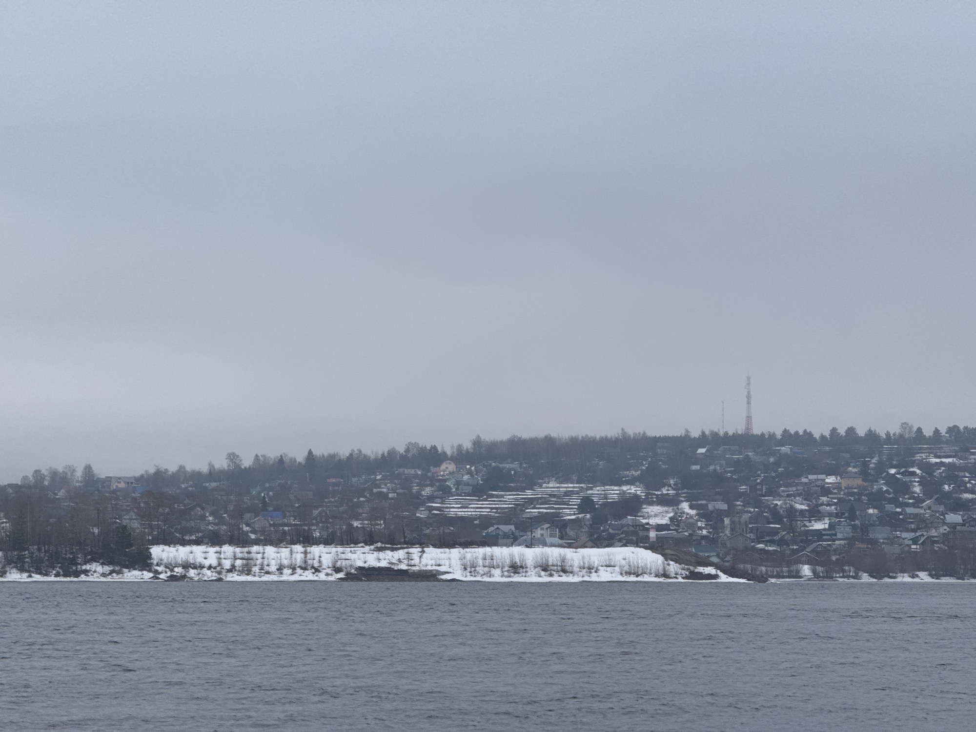 The image shows a waterfront view in Chaikovsky, Russia, on an overcast winter day. The foreground is dominated by a body of water with gentle ripples. Beyond the water is a snow-covered shoreline with bare trees and shrubs. A dense cluster of residential buildings ascends the hillside behind the shoreline, with a mix of single-family homes and multi-story structures. Many of the buildings are also covered in snow. A tall transmission tower is visible amidst the buildings and trees on the hilltop. The sky is uniformly gray and overcast, contributing to the muted, wintry atmosphere. No people are visible in the scene.