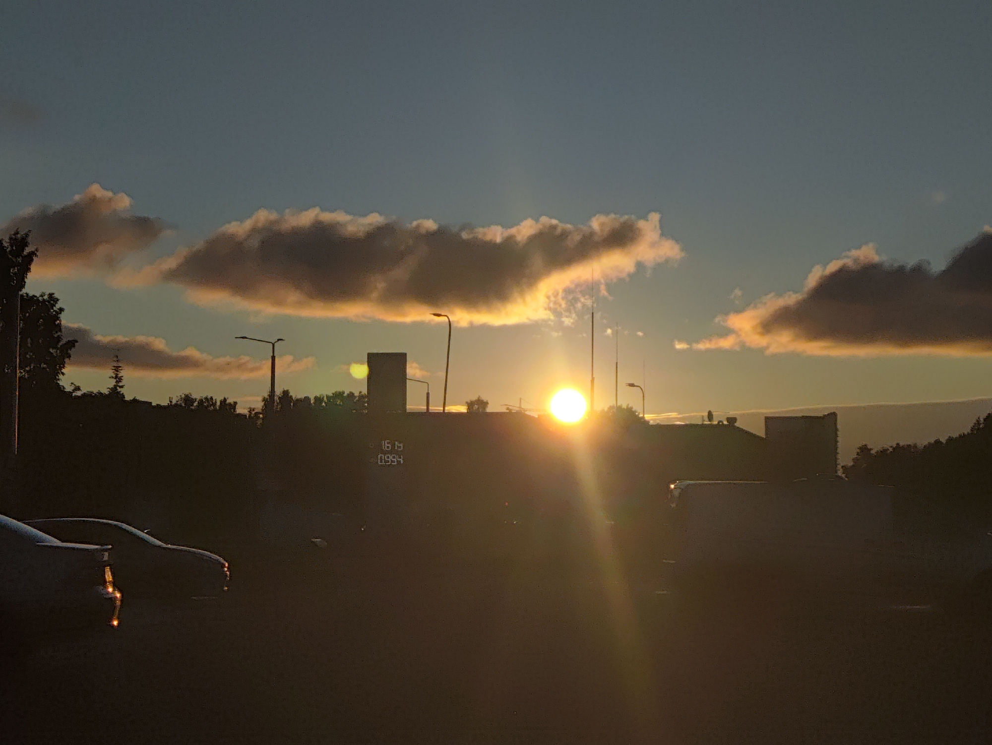The image captures a sunset scene in Tallinn, Estonia. The sun, a bright orb, is low on the horizon, casting a warm glow through scattered clouds. Silhouetted against the vibrant sky are buildings and trees, suggesting an urban or suburban environment. A digital display with numbers "16 15" and "0.994" is visible on one of the buildings, possibly indicating a time or price. In the foreground, the dark silhouettes of cars suggest a parking lot or street. The overall atmosphere is one of twilight tranquility.