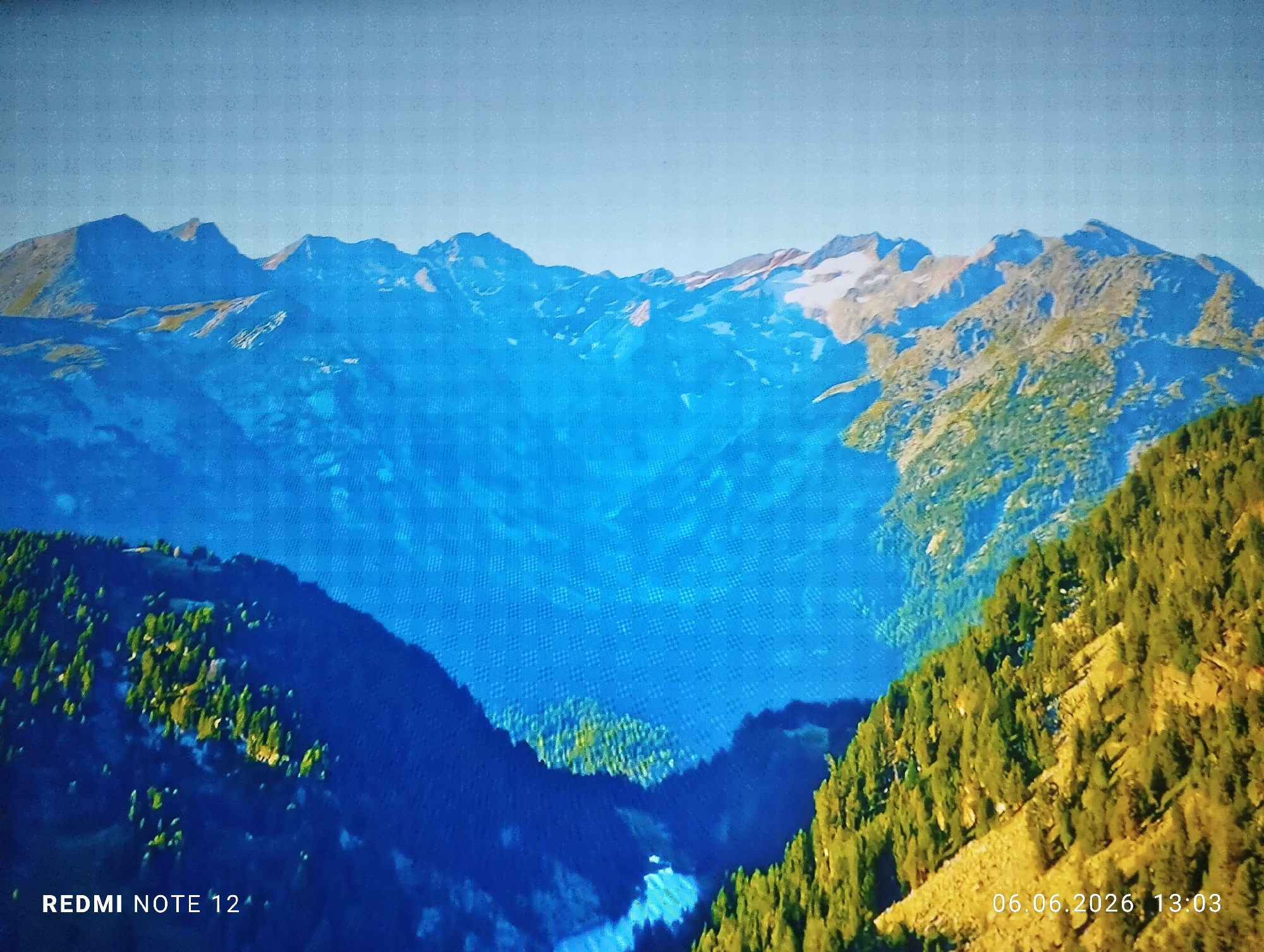 The image displays a layered mountainous landscape, appearing to be a photograph of a digital screen due to a visible grid pattern present across the blue areas. In the foreground, steep slopes covered with dense evergreen trees, characterized by dark green and some yellowish-green hues, ascend from the left and right sides, framing a deep valley in the center. A small, lighter blue/white area at the very bottom center of the valley suggests a river or reflective surface.
The mid-ground is dominated by a series of mountains rendered in a highly saturated, deep blue color, indicating significant atmospheric depth. Beyond these, the background reveals a range of higher mountain peaks under a clear, pale blue sky. Several of these distant peaks show distinct patches of white, indicative of snow or glaciers. No people, animals, or man-made structures are visible within the natural landscape.
Text overlays are present in the bottom corners of the image: "REDMI NOTE 12" is visible in white font in the bottom left, and "06.06.2026 13:03" is visible in white font in the bottom right.
The landscape depicted, characterized by high, snow-capped mountains and alpine forests, is inconsistent with the coastal, low-lying topography of Burgas, Bulgaria. Therefore, this image does not depict a location in Burgas, Bulgaria.