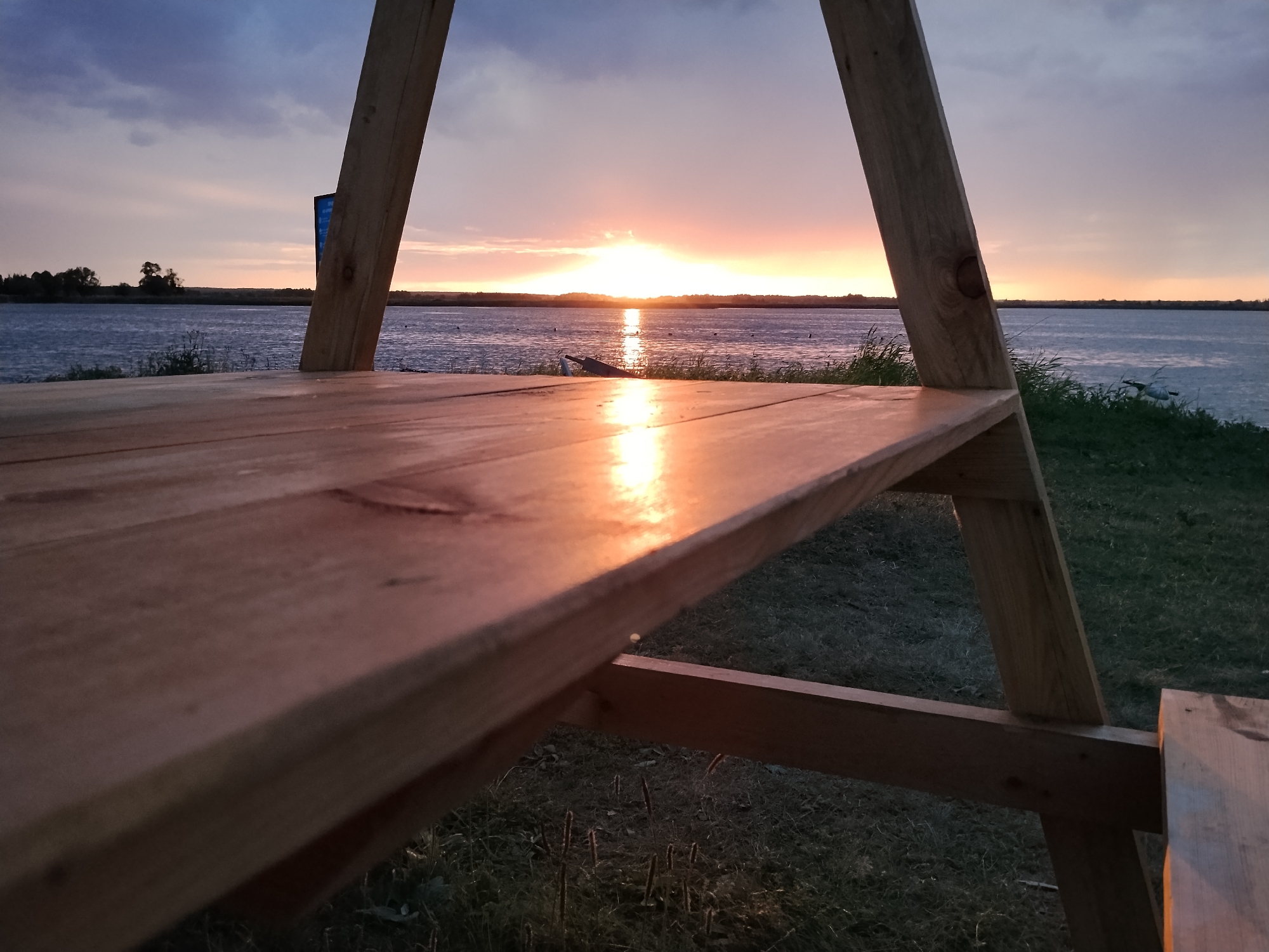 A low-angle shot captures a serene sunset over a body of water, viewed through the frame of a wooden structure, possibly a picnic table or bench. The sun, a bright orb on the horizon, casts a warm, golden glow across the water and the foreground wooden surface, highlighting its texture. The sky transitions from a soft pink and orange near the sun to a deeper blue with scattered clouds higher up. The water is calm, with a gentle ripple reflecting the sky's colors. In the distance, a silhouetted tree line marks the far shore, with some distant structures barely visible. A small boat or kayak is positioned on the water, adding a touch of human presence to the scene. The immediate foreground is a grassy area, with blades of grass catching the fading light. The overall mood is peaceful and tranquil, evoking a sense of quiet contemplation as day turns to night. The time of day is clearly sunset. There is no visible text in the image.