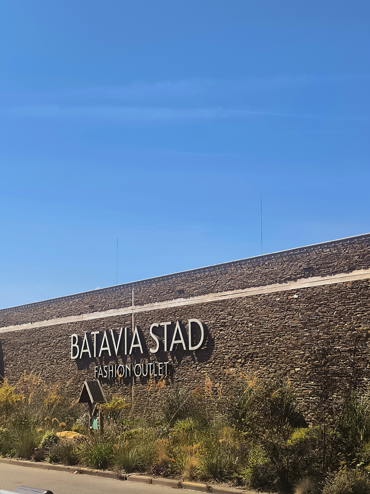 A daytime, eye-level, medium shot captures the exterior of a Batavia Stad Fashion Outlet building in Lelystad, Netherlands. The building's facade is constructed of dark, irregular stones and features a prominent white sign that reads "BATAVIA STAD FASHION OUTLET." Below the sign, a small, dark wooden directional sign stands amidst lush green and brown foliage and grasses. The sky above is a clear, bright blue with a few thin, wispy white clouds. A thin, dark vertical antenna extends from the roofline of the building towards the sky. The foreground shows a portion of a paved road and sidewalk.