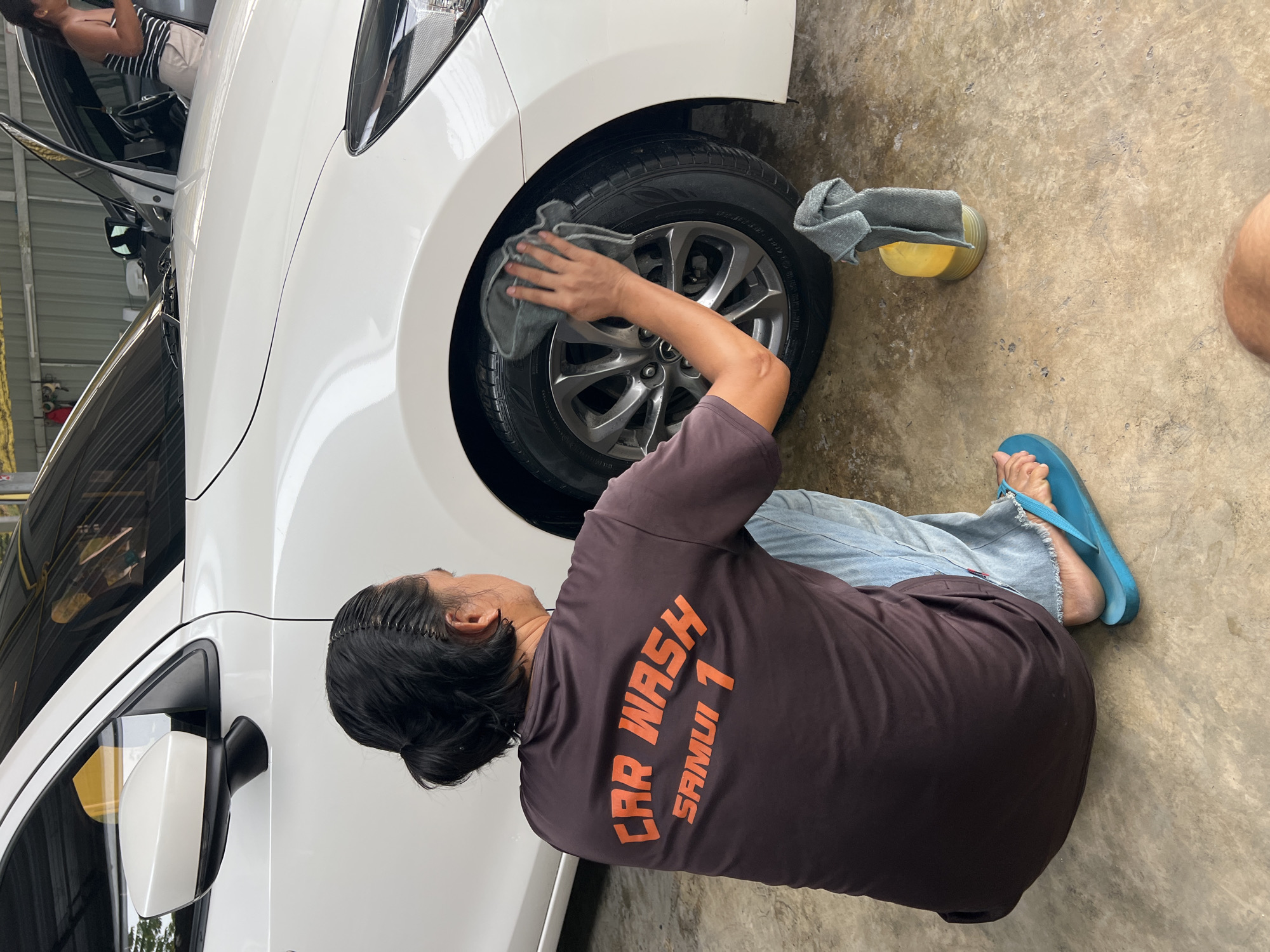 A person wearing a brown t-shirt with "CAR WASH SAMUI 1" printed in orange and light blue jeans is kneeling on a concrete floor and wiping the tire of a white car with a gray microfiber cloth. A yellow bottle with a gray microfiber cloth draped over it stands on the floor near the tire. The scene is outdoors, likely in a car wash facility, indicated by the presence of a corrugated metal roof structure in the background. The overall environment appears to be a somewhat utilitarian setting. The person's feet are clad in blue flip-flops. In the background, another person is visible standing near a car with its door open.