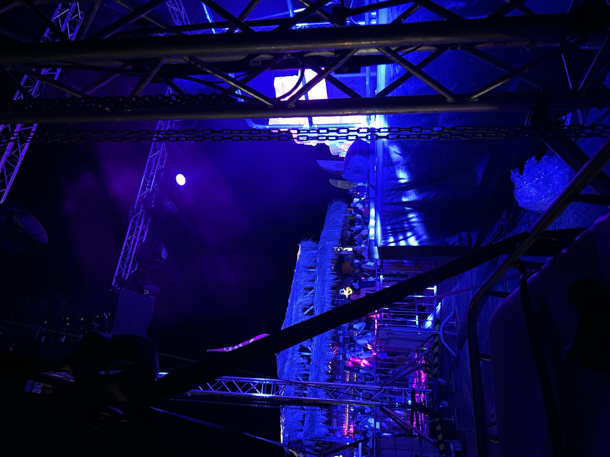 The image depicts a nighttime outdoor scene at a festival or event in Sozopol, Bulgaria. The foreground is dominated by dark metal scaffolding and chains, suggesting a stage or production area. In the midground, a crowded area with a thatched-roof structure, possibly a bar or food stall, is illuminated by vibrant purple and blue lights. People are visible gathered around this structure and in front of it, interacting and enjoying the event. The background shows a dark sky with more stage lighting, including a prominent bright white light on a stage in the distance. The overall atmosphere is lively and energetic, characteristic of an outdoor music or entertainment event.