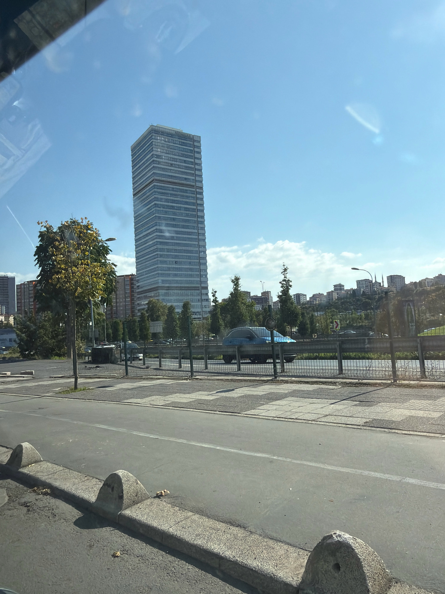The image shows a daytime view of a street in Ataşehir, Turkey. A prominent skyscraper with a glass facade is visible in the background, along with other buildings and trees. A blue car is parked on the side of the road, and there is a pedestrian crossing in the foreground. The sky is clear and blue with some scattered clouds. The street is paved with asphalt, and there are concrete barriers along the side of the road. The scene appears to be a typical urban environment.
