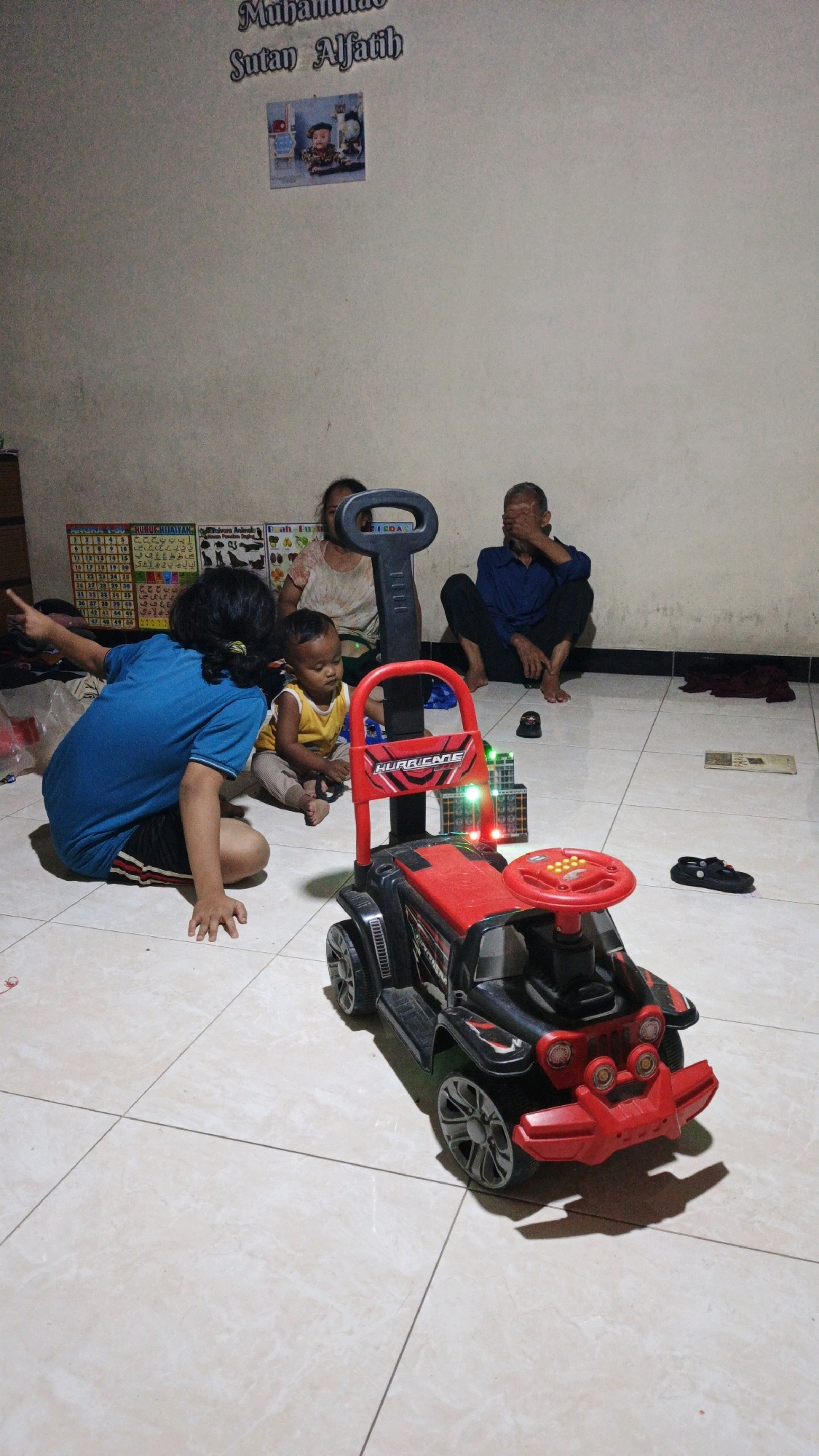 In a dimly lit room with tiled flooring, likely in Bono, Indonesia, four individuals are present. In the foreground, a black and red toy jeep with a push handle is centrally positioned. To the left of the toy jeep, a child wearing a blue t-shirt and dark shorts sits on the floor, facing away from the camera and pointing to the left. To the right of the child and in front of the toy jeep, a baby in a yellow tank top and light-colored pants is seated on the floor, looking down. Behind them, an adult, possibly female, wearing a light-colored top, sits on the floor. Further to the right, an adult male in a dark blue shirt sits on the floor, covering his eyes with his hand. The room's wall in the background displays text and a framed picture. Educational charts are also visible on the wall. Several dark-colored objects, including shoes and fabric, are scattered on the floor around the individuals.