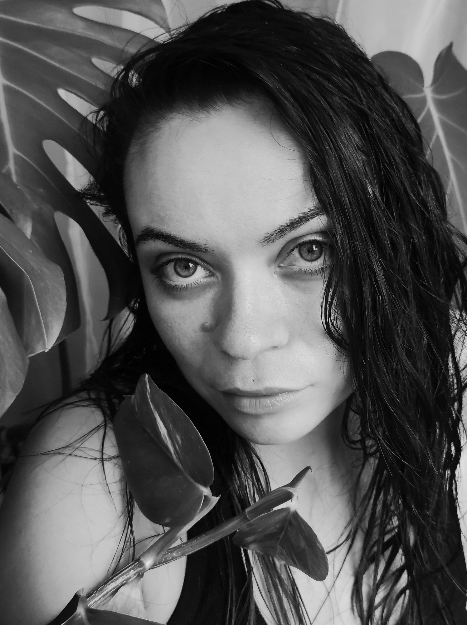 The image is a black and white, close-up portrait of a young woman. She has dark, wet hair that is partially obscuring her face. Her eyes are open and looking directly at the camera. There are large, leafy plants behind her, suggesting an indoor setting with foliage. A vine with leaves is held in front of her face, partially obscuring her features. The lighting is dramatic, creating shadows and highlighting her facial features. There is no indication of the city of Chaikovsky, Russia, or any specific location. The image focuses solely on the woman and the surrounding plants.