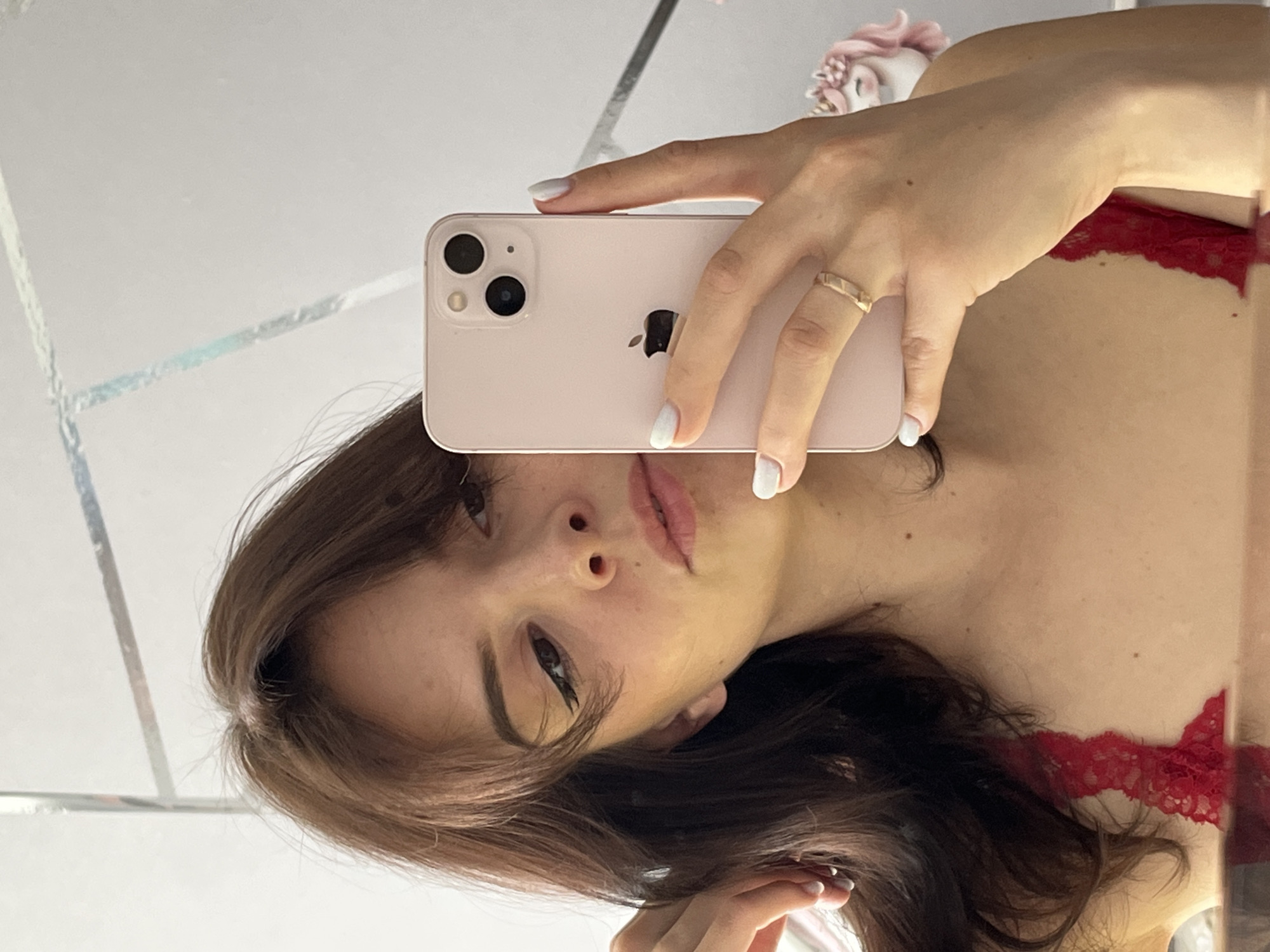 A young woman is taking a selfie in a mirror with a pink iPhone. She has light brown hair, fair skin, and is wearing a red lace top. Her nails are painted white and she has a gold ring on her finger. The background appears to be a room with a mirror on the wall. There is no information to confirm the city of Shushary, Russia as the location.
