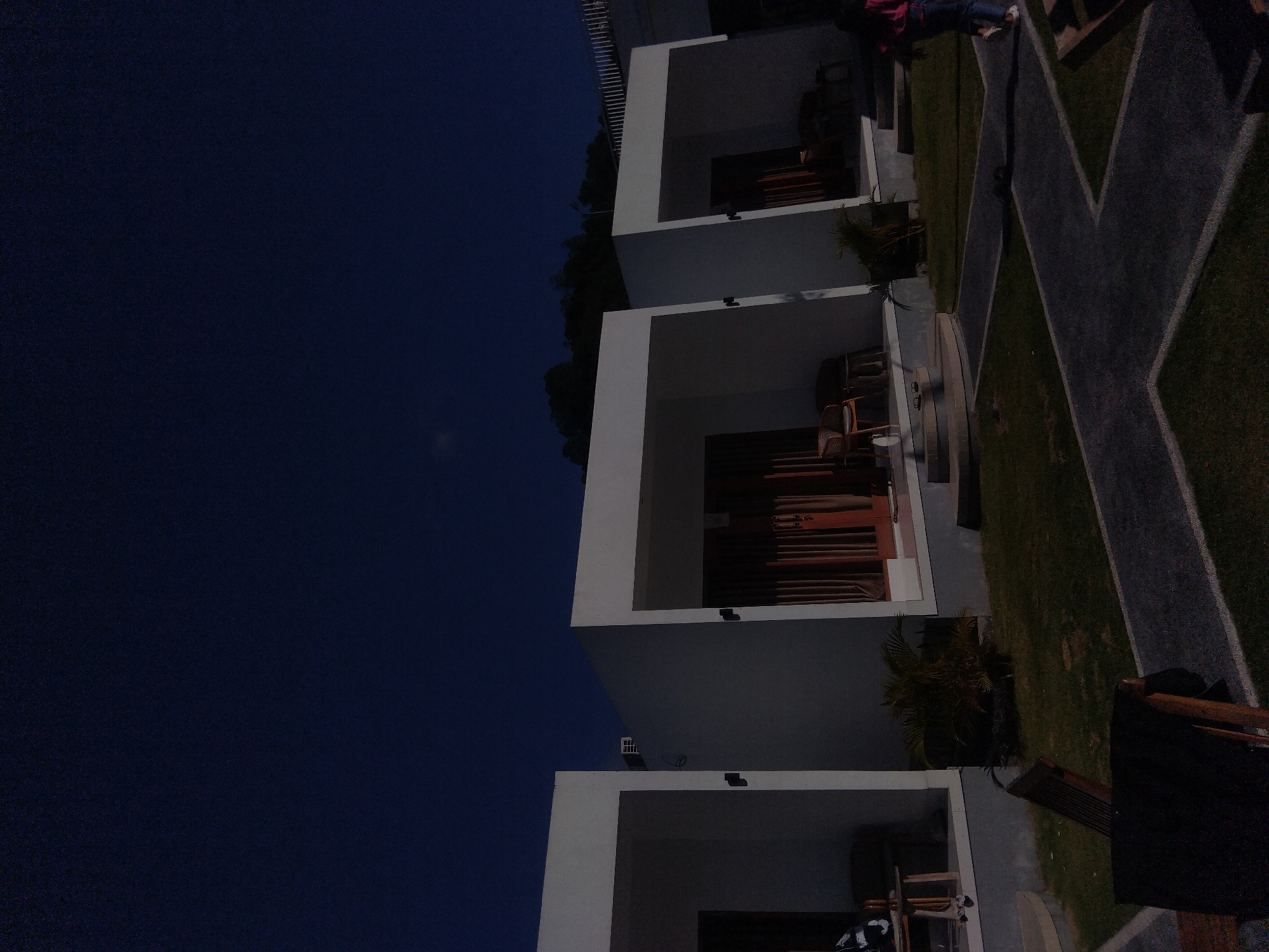 Here's a description of the image:
The image depicts a three-story building with open-air balconies, set against a dark blue sky. The building has a modern architectural style with clean, white lines. Each balcony features furniture. The surrounding area includes grass, a paved walkway with a 'X' pattern. The location appears to be in a city.