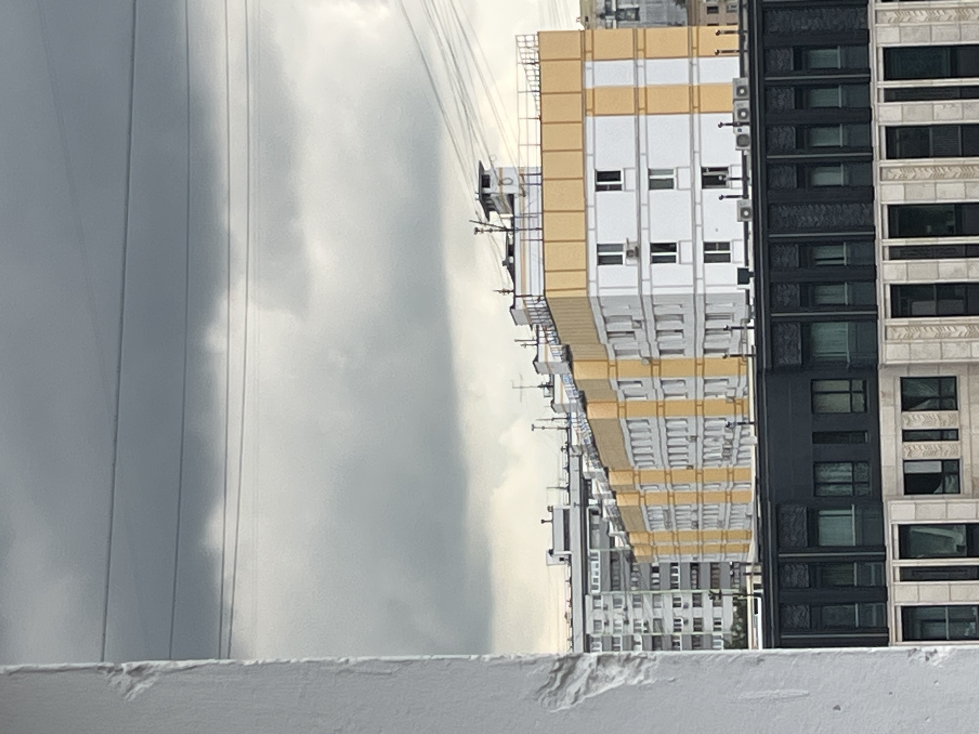 The image depicts a cityscape under a cloudy sky, focusing on several apartment buildings. The foreground on the left shows a textured, light-colored wall, possibly a balcony or building edge. Beyond this, a row of multi-story apartment buildings is visible. The most prominent building in the middle ground is light-colored with yellow vertical accents and white horizontal bands. It has numerous windows and air conditioning units mounted on its exterior. Behind this building, another taller apartment building with a grey and white facade is visible. In the background, more buildings and overhead power lines are apparent against the overcast sky. The scene suggests an urban environment in Moscow, Russia, characterized by residential architecture. There are no people or vehicles visible in the frame. The lighting is diffused due to the cloud cover, creating a muted color palette.