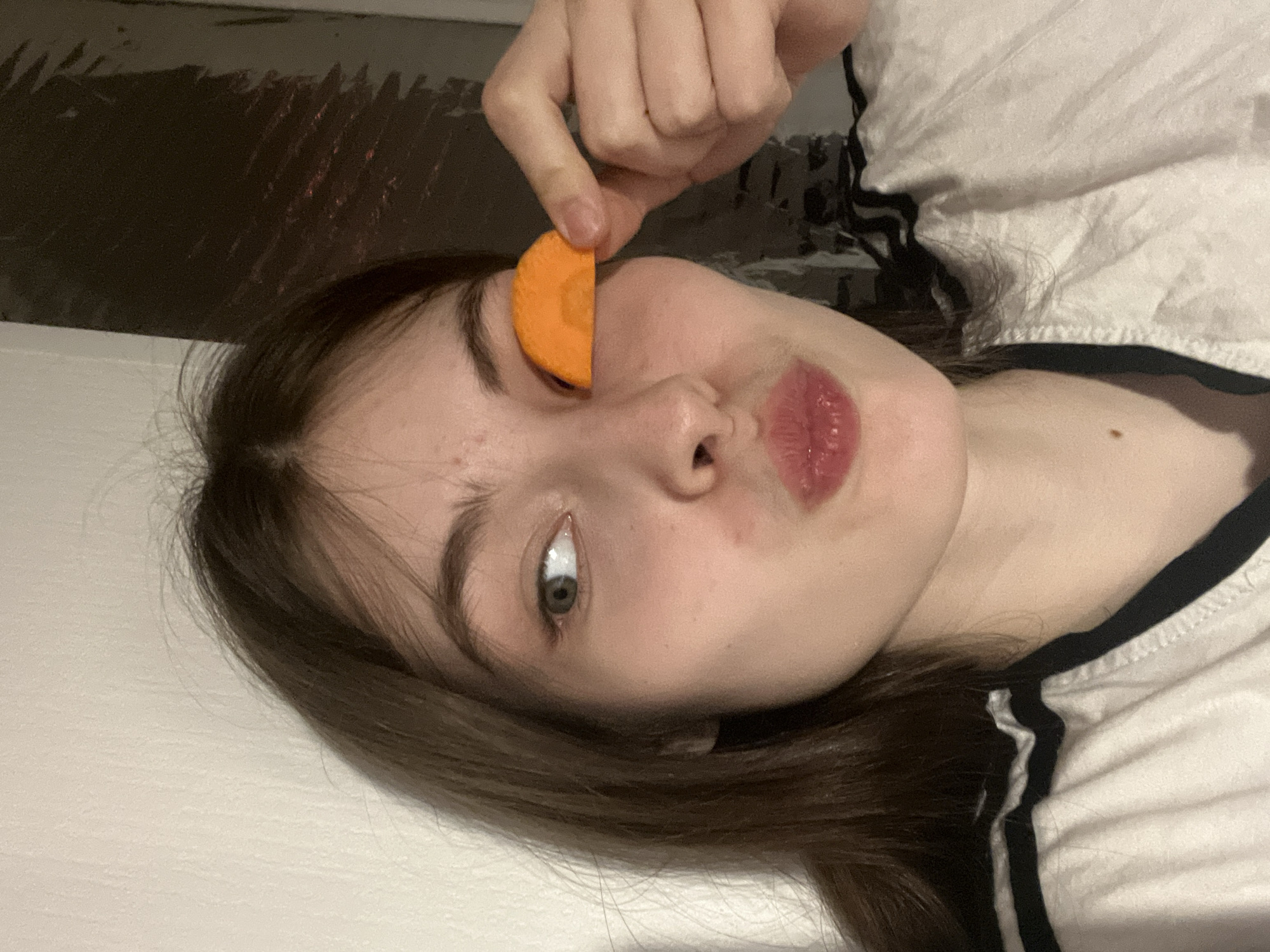 A young woman is holding a slice of carrot over her left eye. She is lying on a white surface, possibly a bed, wearing a white t-shirt with black trim around the collar and shoulders. Her dark hair is parted to the side and her lips are slightly pursed. The background appears to be a wall or surface with a dark, possibly patterned covering. The location is not discernible from the image, and it is not possible to determine whether it is in Saint Petersburg, Russia.