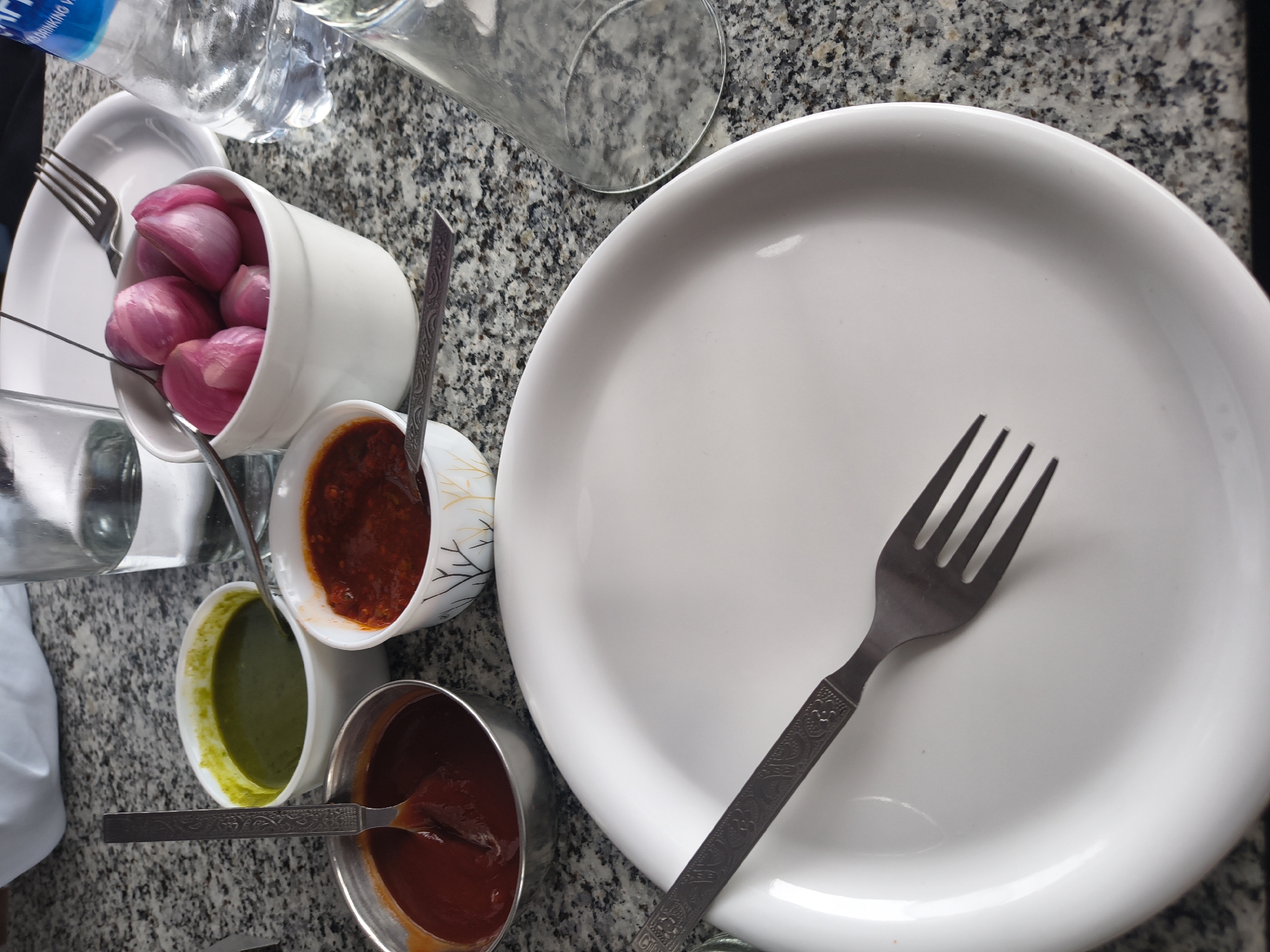 This is a close-up, high-angle, indoor shot of a dining table at a restaurant in Dharamshala, India. The table has a speckled granite surface. In the foreground, a white plate is placed centrally, with a metal fork resting on it. To the left of the plate, there are three small bowls containing various food items. The leftmost bowl, made of metal, holds a dark red sauce. Next to it, a white ceramic bowl contains a bright green chutney. To the right of these, another white ceramic bowl, decorated with a subtle black leaf pattern, holds a reddish-orange chutney or sauce. Behind these bowls, a white ceramic bowl is filled with quartered red onions. Several glasses, one partially filled with water and another empty, are visible on the table, along with a plastic bottle of water with a blue label. A metal spoon is also present, partially submerged in one of the food bowls. The lighting suggests an indoor setting, possibly with natural light from a window.