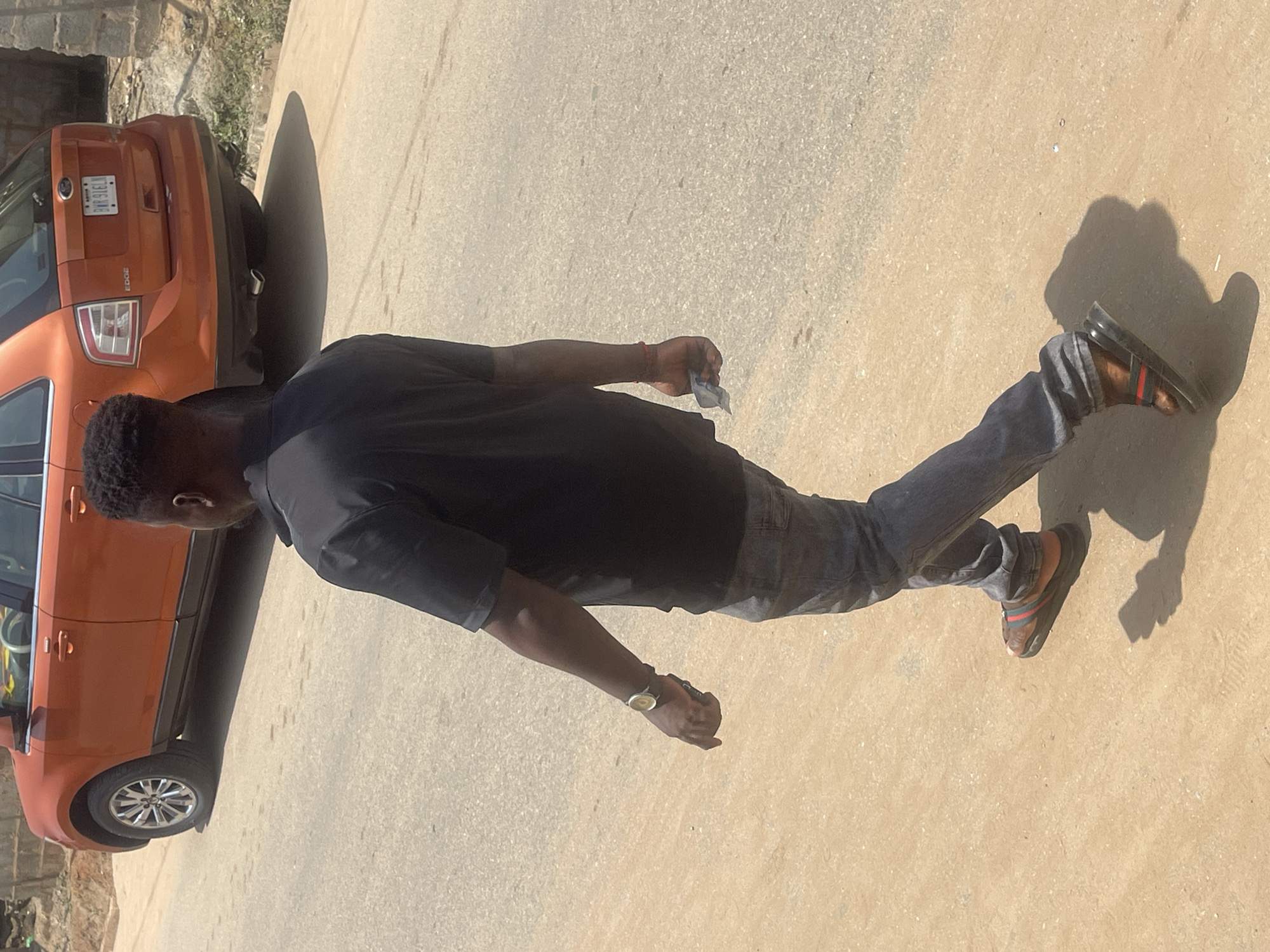 In Abuja, Nigeria, a man is captured from behind, walking away from the camera on a dusty asphalt road. He is wearing a black t-shirt, grey jeans, and dark sandals with red and green stripes. On his left wrist, he wears a wristwatch with a gold-colored band and a white face. In his right hand, he holds a small, folded piece of paper.
In the background, an orange Ford Edge SUV is parked on the side of the road. The vehicle has a license plate with the characters "BWR 91ELN." The environment appears to be an urban or semi-urban area, characterized by the road surface, the parked vehicle, and some background structures that suggest a developing area, possibly with some unpaved ground and vegetation. The lighting suggests it is daytime, with distinct shadows cast by the man and the vehicle.