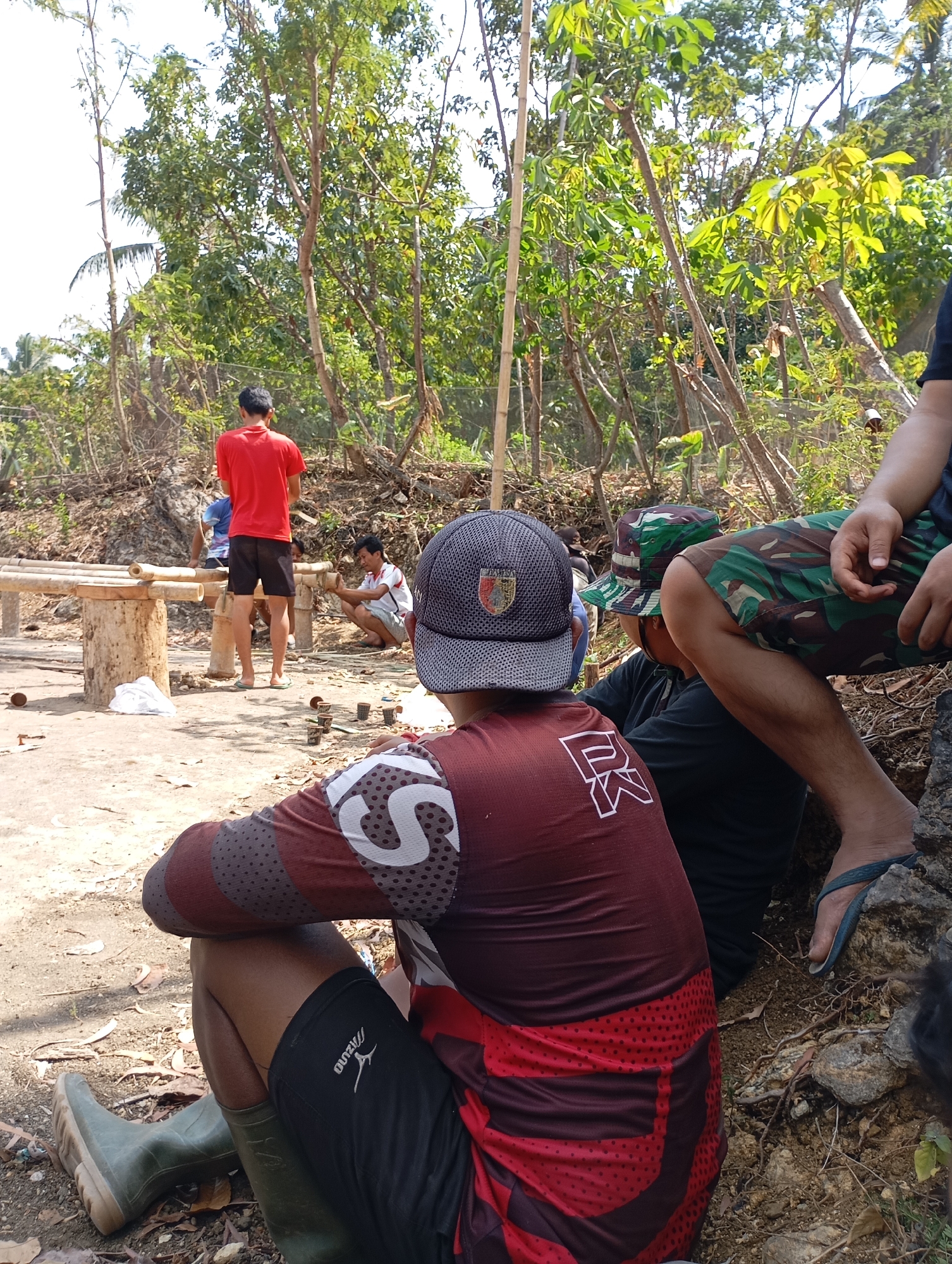 The image depicts an outdoor scene in the vicinity of Ketepung, Indonesia, with several individuals gathered. In the foreground, a person wearing a maroon and black patterned long-sleeved shirt and black shorts is seated, facing away from the camera. This individual also wears green rubber boots. To the right of the foreground subject, another person, partially visible, is seated and wearing camouflage shorts and a dark t-shirt. This person's legs are crossed and they are wearing blue flip-flops. In the middle ground, a person in a red t-shirt and black shorts stands near a bamboo structure. To their left, another individual is seated. Further back, a person wearing a camouflage hat and shorts is seen. The environment is characterized by dry, rocky terrain with scattered trees and vegetation. A low bamboo bench-like structure is present in the mid-ground, and several small cups or containers are arranged on the ground. The lighting suggests a sunny day.