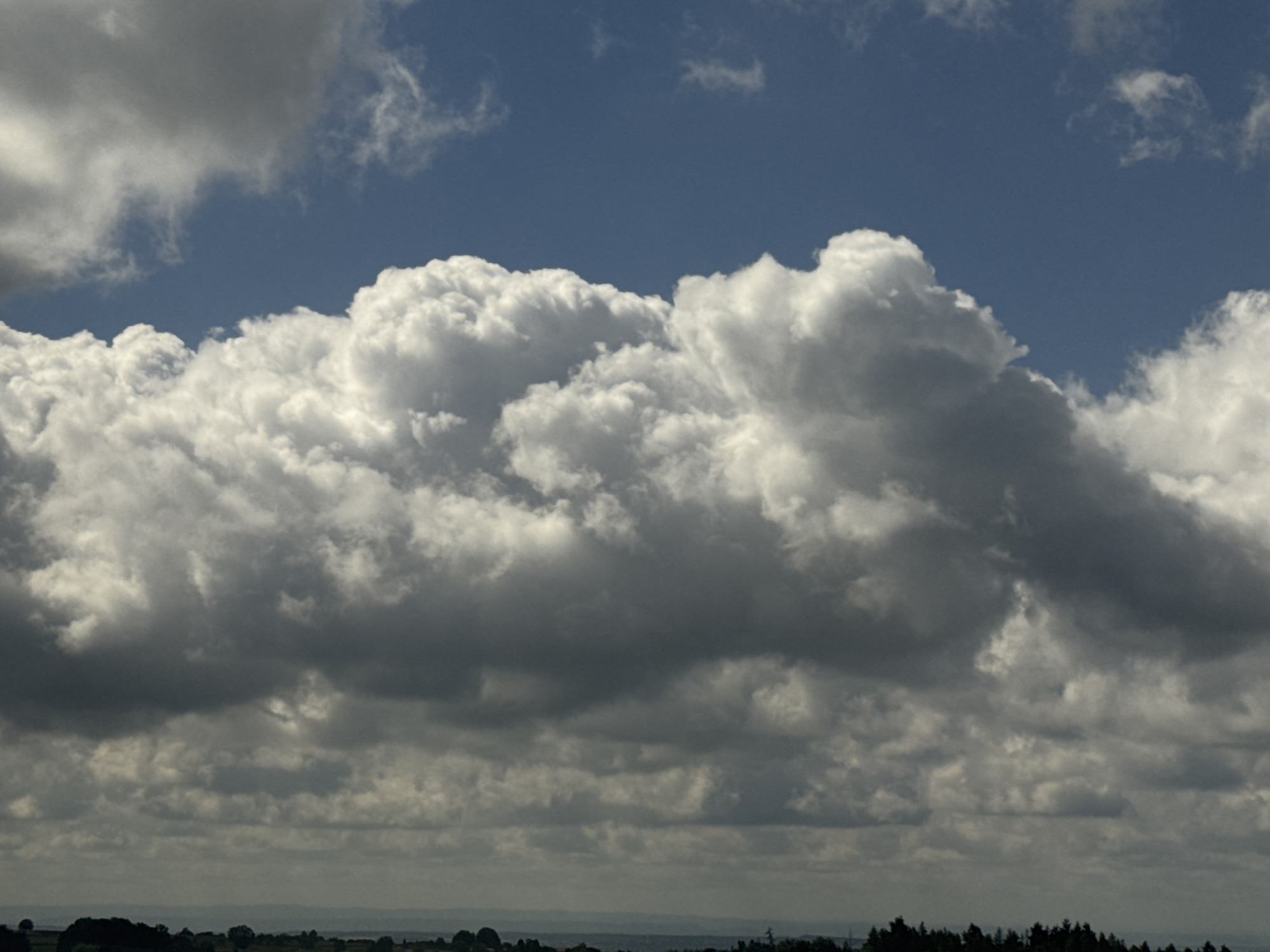The image depicts a wide-angle view of a cloudy sky above a distant, dark landscape of trees and hills. The sky is a dominant feature, filled with a variety of clouds. In the upper portion of the image, there are areas of bright white, puffy cumulus clouds contrasted against a deep blue sky. Lower down, the clouds become denser and darker, with shades of gray and white, suggesting a build-up of storm clouds or a change in weather. The bottom of the image shows a dark, silhouetted line of trees and rolling hills, indicating a rural or natural environment, possibly on the outskirts of Strümpfelbrunn, Germany. There are no people or discernible objects in the immediate foreground or mid-ground, making the sky and the distant landscape the sole subjects of the photograph. The lighting suggests it is daytime, with the sun likely behind or to the side of the clouds, creating dramatic contrasts in brightness.