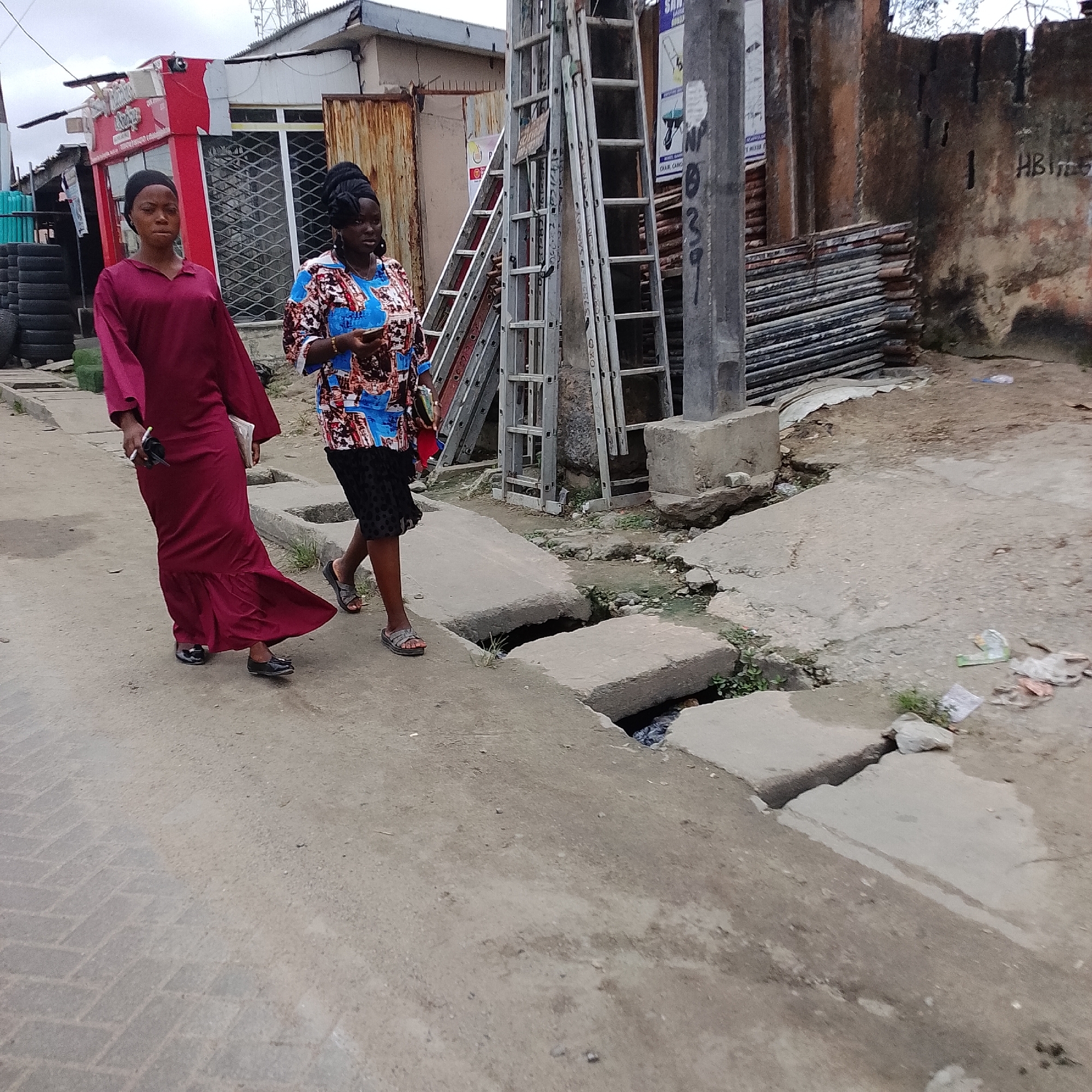 Two women walk along a dirt and paved path in Ado, Nigeria. The woman on the left wears a long, flowing maroon dress and black shoes. The woman on the right wears a patterned shirt and a black skirt with polka dots, along with sandals. They appear to be walking side by side, with the woman on the right looking towards her phone. The environment is urban, with buildings and a damaged drainage system visible. In the background, there are structures that appear to be shops, stacks of metal sheeting, and ladders leaning against a pole. Tires are also visible to the left of the frame. The ground is a mixture of concrete and dirt, with some vegetation growing in the crevices of the damaged drainage. Litter is scattered along the path. The sky is overcast.