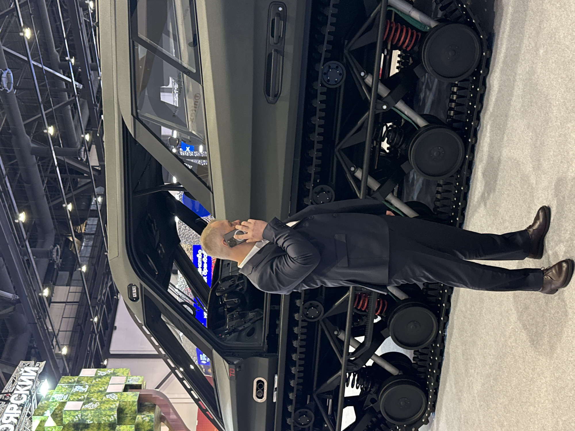 An indoor scene depicts a man in a dark suit and light-colored shirt standing next to a large, olive-green tracked vehicle. He is engaged in a phone call, holding a light-colored smartphone to his right ear. The man has short, light-colored hair and is positioned slightly to the left of the vehicle's opened door.
The vehicle is a prominent subject, showcasing its robust, exposed track system with multiple black road wheels and red suspension springs. The cabin features dark-tinted windows, a visible door handle, and internal controls.
The setting strongly suggests an exhibition or trade show, likely in Pushkin, Russia. The background reveals a high ceiling with industrial-style piping, ventilation ducts, and numerous light fixtures. To the left, a display made of interconnected green cubic elements is partially visible. The floor is covered in a light-colored carpet or exhibition flooring.
Visible text includes the Cyrillic word "ОЯРСКИЙ" (OYaRSKIY) on the green display. Faint text, possibly "Quinox," can be discerned on the vehicle's window, and a red "T8" is visible on the vehicle's door frame near the dashboard. The man appears focused on his conversation, amidst what seems to be a display of specialized vehicles.