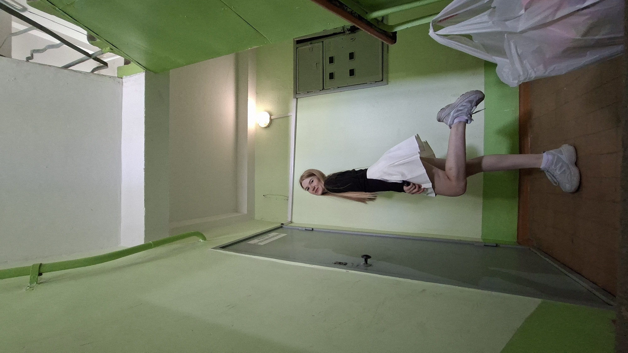 In the city of Chaikovsky, Russia, within a dimly lit hallway or stairwell, a young woman stands near a door. She has blonde hair and is wearing a black long-sleeved top and a white pleated skirt. Her right leg is bent and raised behind her, and she is looking back at the camera with a slight smile. To her left is a grey metal door with a handle and what appears to be a lock mechanism. The walls of the hallway are painted a light green, with a darker green stripe near the floor. A green pipe runs along the wall near the ceiling. Further down the hallway, a metal electrical panel is mounted on the wall. A light fixture is visible on the ceiling. To the right of the woman, a portion of a staircase with a green railing is visible, and a large white plastic bag filled with items rests on the floor. The floor is covered with brown linoleum.