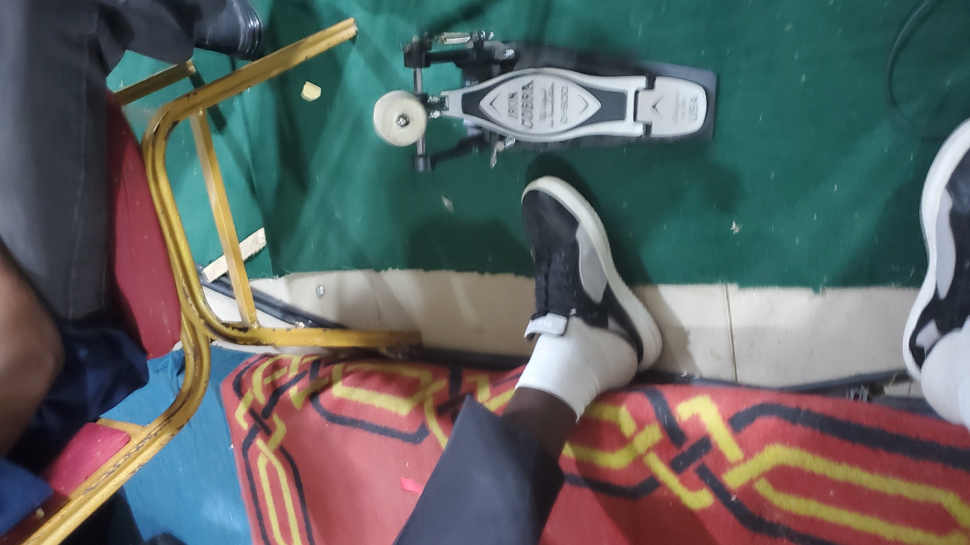 The image depicts a close-up of a person's legs and feet, one of which is resting on a bass drum pedal. The scene appears to be indoors, possibly a rehearsal space or a performance venue, given the presence of the drum pedal and a chair. The person is wearing dark pants, white socks, and black and gray sneakers. The bass drum pedal is a black and silver "Iron Cobra" model. A red and yellow patterned rug is visible to the left of the frame, and a green surface, likely carpet or a floor covering, is to the right. The lighting is dim, casting shadows that suggest an indoor environment. The overall impression is of someone preparing to play a musical instrument, specifically drums, in a casually set-up space. The location in Jalingo, Nigeria, is not discernible from the visual information presented in the image.