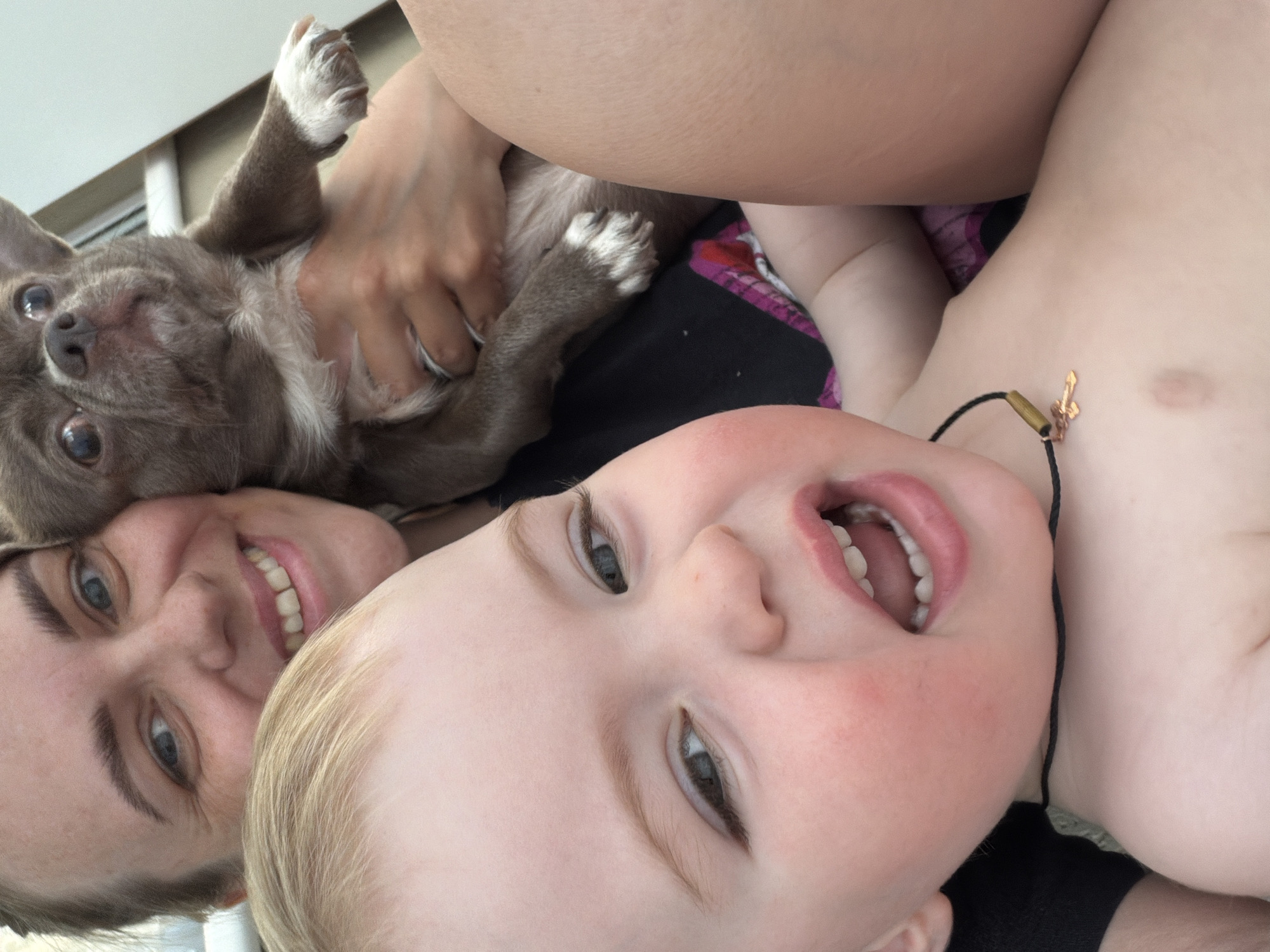 The image presents a close-up, tilted view of a fair-skinned adult woman with short blonde hair smiling, a young child with light blonde hair lying on their back with an open mouth and visible teeth, and a light grey-brown dog with white paws being held and positioned closely to the woman's face. The child is wearing a black cord necklace with a gold-colored pendant, and the woman's hand is partially visible, holding the dog. The background is slightly out of focus and indistinct, and it is impossible to identify the location of the city of Votkinsk in Russia.