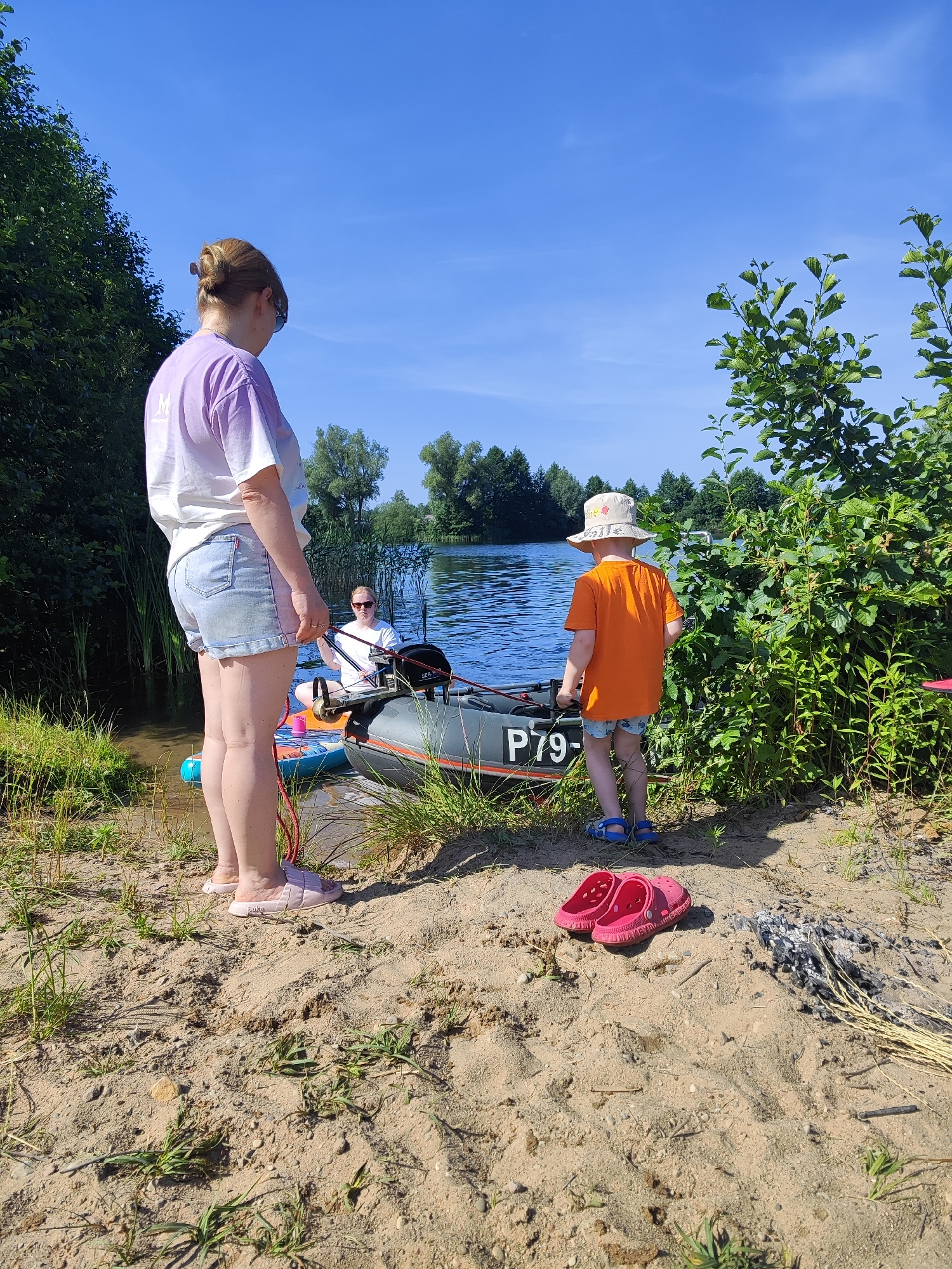 In the city of Rovnoe, Russia, on a sunny day, a sandy shoreline meets the calm waters of a lake. A woman, wearing a light purple and white tie-dye t-shirt and denim shorts, stands on the sand, facing away from the camera, her hair in a bun. Beside her, a young child in an orange t-shirt and a patterned sun hat, is looking at a gray inflatable boat beached on the shore. The boat has "P79-" visible on its side. Another person, wearing a white t-shirt and sunglasses, is seated in the boat. Two pink flip-flops lie on the sand near the child, and a small pile of ashes and dry grass is visible further to the right. The background is filled with lush green trees and a clear blue sky.