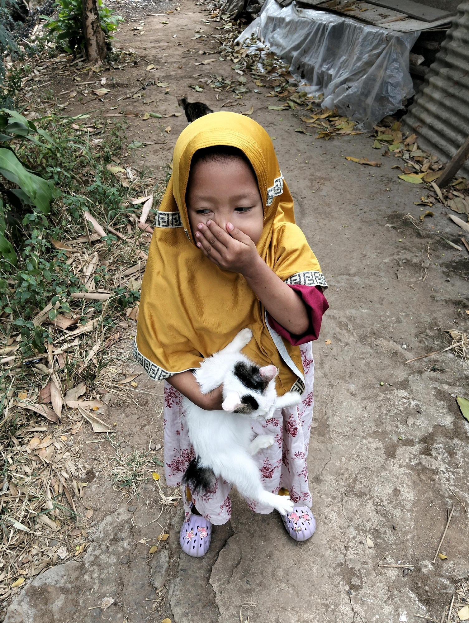 A young child, wearing a yellow hijab and a floral patterned dress, is holding a white and black cat. The child's right hand is covering their mouth, and their head is tilted downwards. The cat is held close to the child's chest, with its body facing upwards and its head towards the child's right. The child is standing on a dirt path, with dry leaves and sparse vegetation on either side. In the background, there appears to be a corrugated metal structure and some plastic sheeting. The scene is outdoors, with natural lighting. The location is the village of Cileungsi, Indonesia.