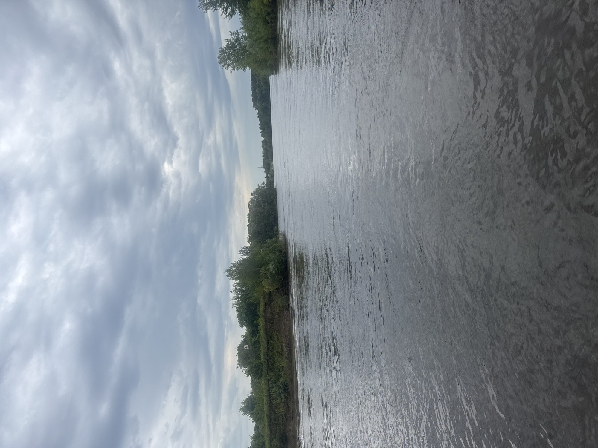 The image depicts a wide river under a cloudy sky. The riverbanks are lined with dense green trees. On the left bank, a small white sign with a red cross is visible among the trees. The water appears calm with some ripples. The overall scene suggests a natural landscape with a hint of human presence. The location is identified as the Belaya River in Ufa, Russia.
