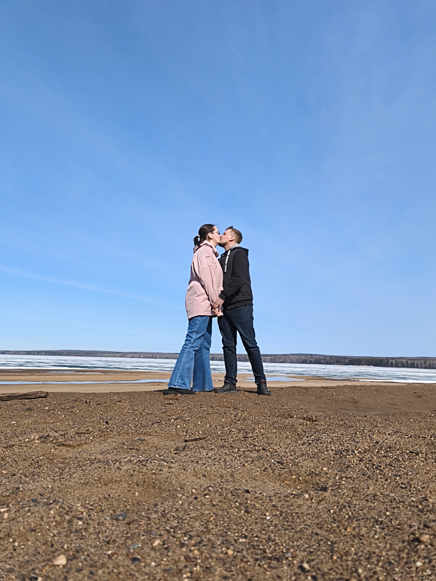 Two individuals, a male and a female, are standing on a sandy shore, kissing. The female is wearing a light pink jacket and blue jeans, while the male is wearing a black jacket and dark blue jeans. They are holding hands. Behind them is a body of water with partially melted ice, and a treeline along the horizon. The sky is blue with some light clouds. The location is in the city of Unknown, Unknown.