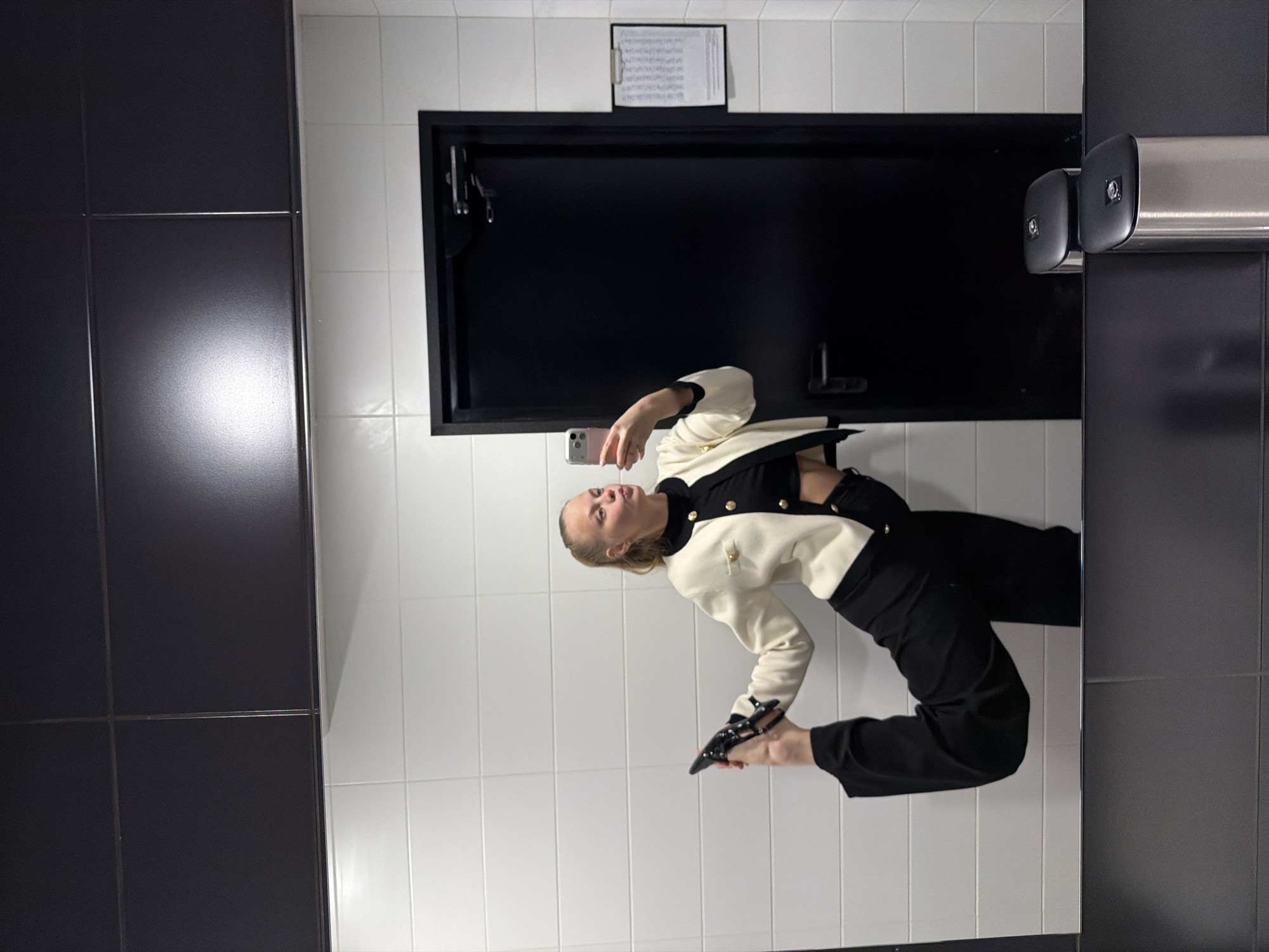 A young woman is captured in a mirror selfie, posing in what appears to be a tiled indoor space. She is looking at her smartphone, which she holds in her left hand to take the photo, and has her mouth slightly agape, suggesting a playful or expressive moment.
The woman is dressed in a stylish cream-colored jacket with black accents, featuring gold buttons. She is also wearing black trousers and holds a black studded shoe in her right hand, while the other shoe is not visible. Her hair is pulled back.
The background consists of white rectangular tiles on the wall where the mirror is located, and dark grey or black square tiles on an adjacent wall or partition to the left. Behind the woman, there is a black framed opening or door. Above this black frame, a clipboard with a printed document is visible, taped to the white tiled wall. The text on the clipboard is too small and blurry to be deciphered. To the right of the frame, there are metallic, cylindrical bars or handles, possibly belonging to another door or partition. The overall setting, with its tiled walls and specific fixtures, suggests a restroom, changing room, or similar public/commercial interior. The lighting is artificial, and the time of day is not discernible. The location context of Izhevsk, Russia, is not visually verifiable from the image itself.