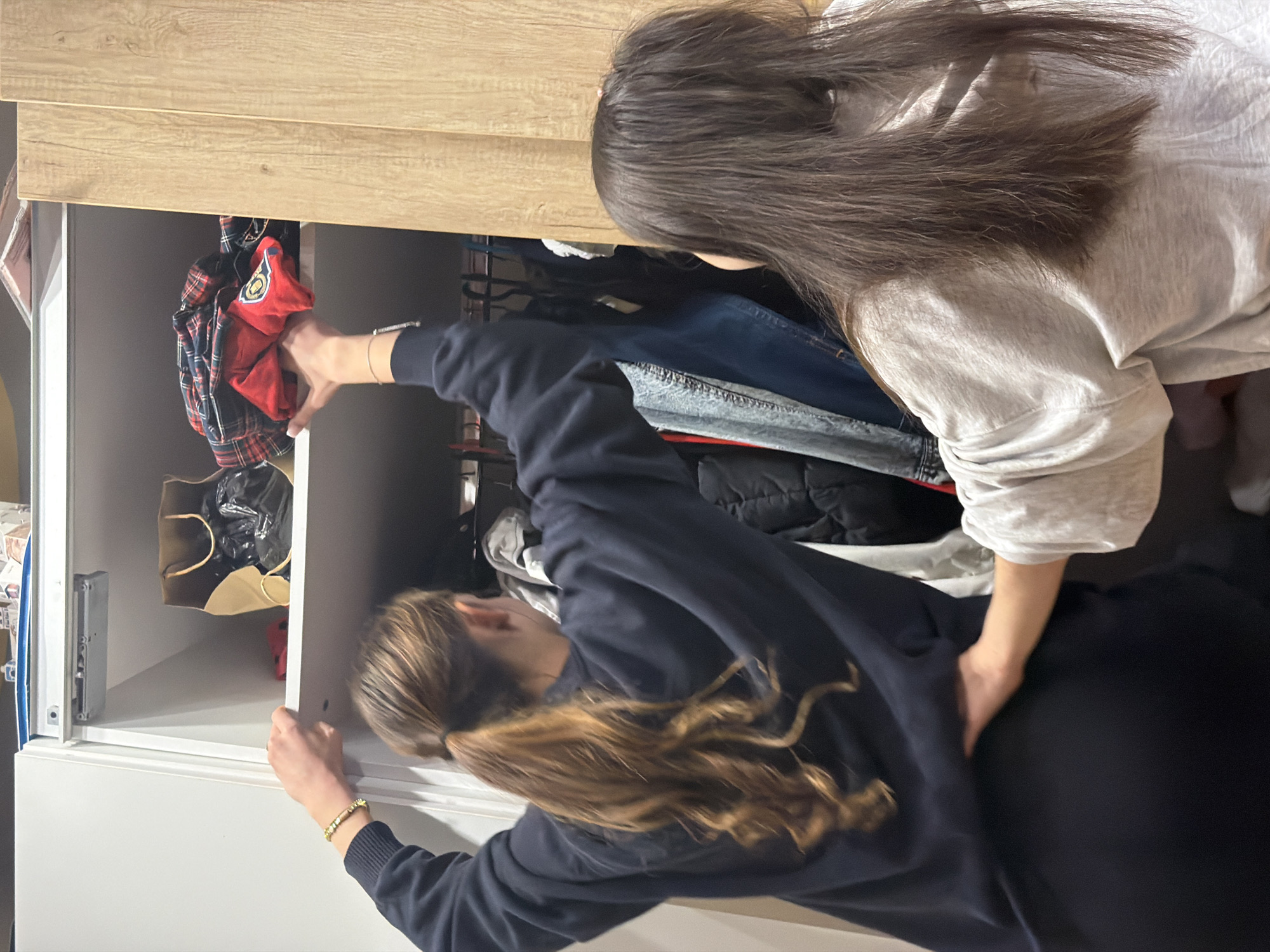The scene depicts two individuals interacting with an open wardrobe inside a residential space in Sofia, Bulgaria. A person with long, wavy brown hair styled in a ponytail, wearing a dark blue long-sleeved top and dark bottoms, stands on the left, reaching upwards with both hands. Their left hand is placed on a white shelf edge, while their right hand is manipulating a folded pile of red and black plaid fabric on the top shelf of the wardrobe. A gold-colored beaded bracelet is visible on the left individual's wrist. To their right, a second individual with long, straight dark brown hair, wearing a light-colored long-sleeved top, has their left hand resting on the lower back/hip area of the first person. The wardrobe features a white left section and a light wood-grained right section. The top shelf contains a brown paper bag holding dark contents, a small red object, and the plaid fabric. Below this shelf, a clothes rail holds various hanging garments, including several denim items and a dark quilted jacket. The setting is indoors, with artificial lighting illuminating the wardrobe's interior.