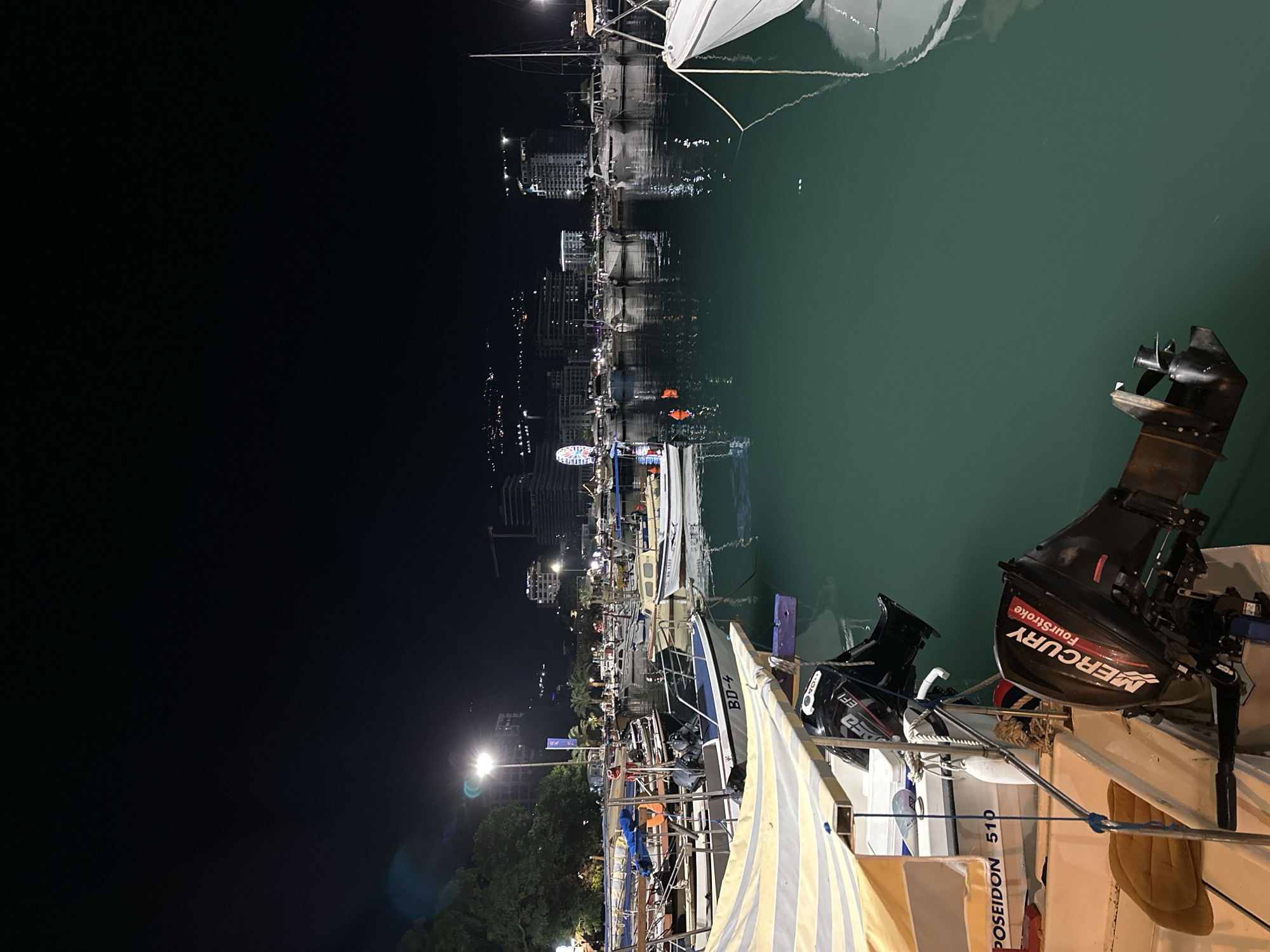 This is a nighttime scene at the marina in Budva, Montenegro. The image is dominated by a dark sky and the illuminated buildings of the city in the background, including a Ferris wheel. The foreground features several boats docked in the water, with their reflections shimmering on the surface. One boat in the immediate foreground has a yellow and white striped awning and a visible "POSEIDON 510" label. Another boat further back shows a "BD-4" marking. Several boats have outboard motors, with one clearly displaying "MERCURY FourStroke". The water appears calm and reflects the lights of the city and the boats. The overall impression is a peaceful night at a coastal marina.
