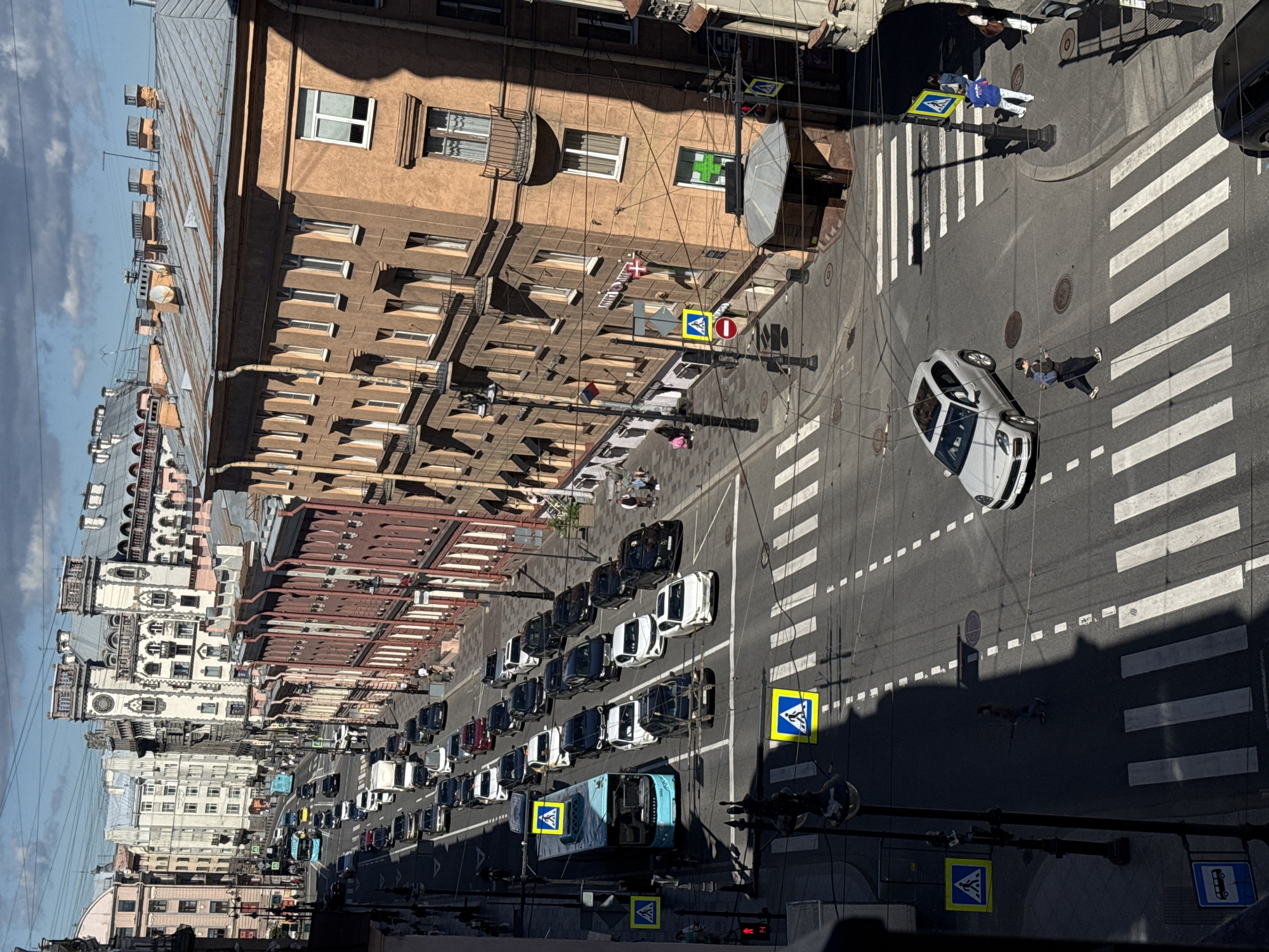 The image depicts an aerial view of a city street, possibly in Saint Petersburg, Russia, given the architectural style of the buildings. The street is lined with multi-story buildings, characterized by ornate facades, balconies, and mansard roofs. Several cars, including a light-colored SUV and a blue bus, are visible on the road. Pedestrians are crossing the street at designated crosswalks. Traffic signals and streetlights are present. The weather appears to be sunny with some clouds in the sky. The street is paved, with white markings indicating lanes and pedestrian crossings. Some of the buildings appear to be commercial, with signs visible on their exteriors. The overall scene is that of a busy urban environment.
