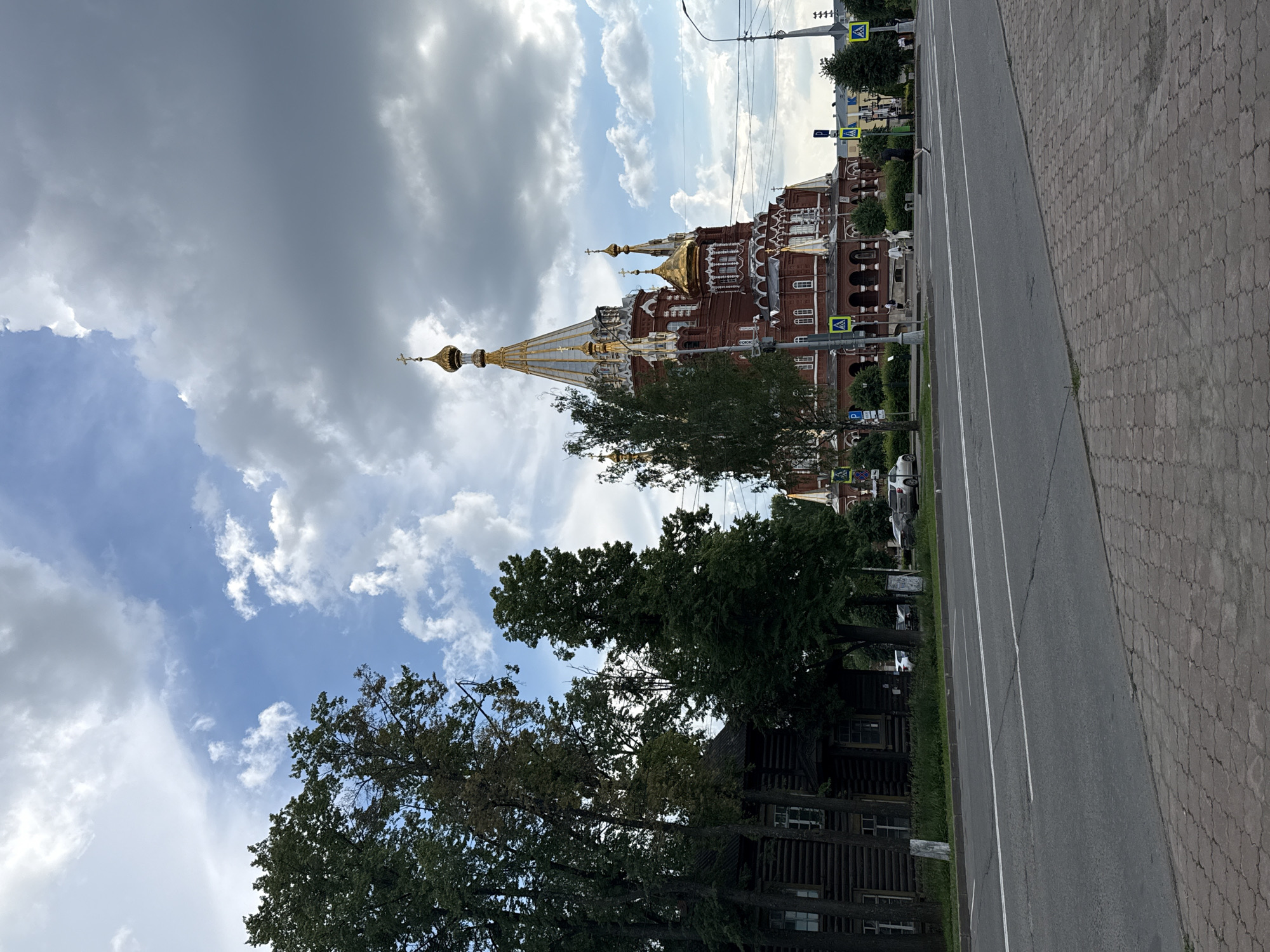 The image shows a view of the Alexander Nevsky Cathedral in Izhevsk, Russia, on a cloudy day. The cathedral is a large, red brick building with golden domes and crosses. To the left of the cathedral is a dark wooden house, and in front of them is a wide road with traffic. There are several trees along the side of the road, and a pedestrian walkway made of brick pavers. The sky is filled with dramatic clouds, with patches of blue sky visible. Several cars are parked or driving on the road. A few people are visible on the sidewalk in front of the cathedral, and one person is running across the street. There are also traffic signs visible, including pedestrian crossing signs and parking signs. The overall impression is of a city scene with historical and religious architecture.