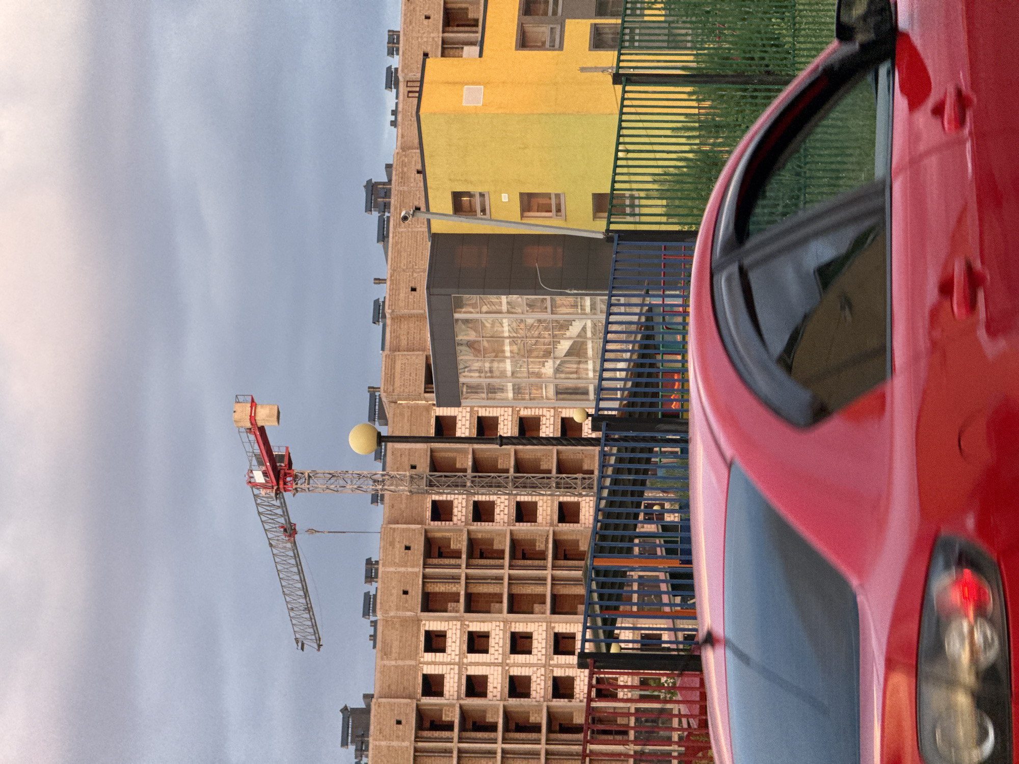 The image depicts an outdoor construction site under a cloudy sky. A prominent red and grey tower crane is visible, positioned above a partially constructed multi-story building made of light-colored bricks and concrete. Adjacent to this building is a brightly painted yellow structure with windows. In the foreground, the rear section of a red car is blurred due to motion, indicating it is either moving or the photo was taken with a slow shutter speed. A dark fence with vertical bars runs across the mid-ground, separating the construction site from a patch of green vegetation behind it. The scene is likely located in Mikhaylovsk, Russia, given the context.