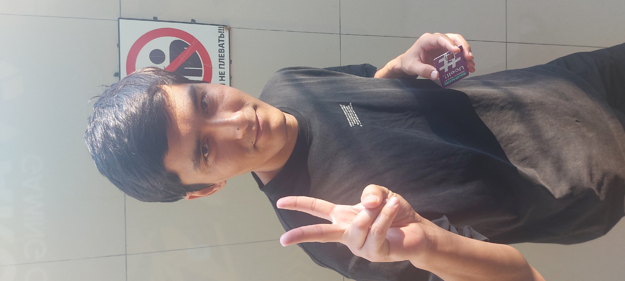 A person is posing outdoors in front of a light-colored wall, likely in Tashkent, Uzbekistan. The person is wearing a dark t-shirt and holding a small, purple and white card with Cyrillic text in their left hand. The card appears to be a prepaid mobile phone scratch card from Ucell, a mobile network operator in Uzbekistan. The person is making a peace sign with their right hand. Behind them is a sign featuring a crossed-out figure spitting, next to the word "Gaming". Below that is the phrase "не плевать!!!" which means "do not spit!!!" The image is oriented sideways.
