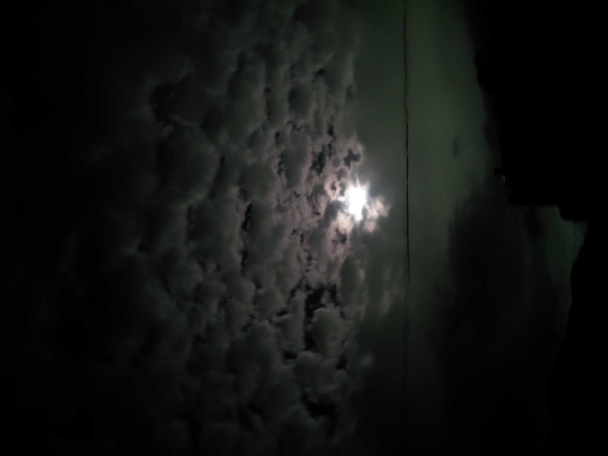 The image captures a dark, cloudy night sky, likely observed from Santa Ana, Venezuela. A powerful, brilliant light source, presumed to be the moon, is partially visible through a dense formation of clouds, dominating the upper right-middle section of the frame. The light strongly illuminates the surrounding dark gray and white clouds, highlighting their varied textures and creating a dramatic contrast against the dark sky. The clouds fill most of the composition, appearing thick and turbulent. On the far right side, a dark, indistinct silhouette, possibly a person's head and shoulder or a part of a nearby structure, is visible against a slightly greenish-tinged dark background. The overall scene conveys a sense of mystery and natural wonder, with the moon's intense glow piercing through the nighttime cloud cover. The time of day is clearly night, with overcast weather conditions. No visible text is present in the image.
