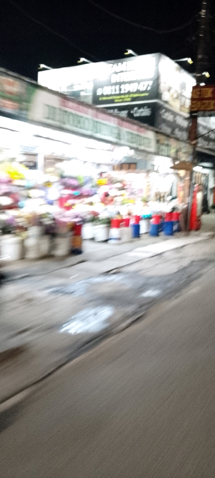 The image shows a street scene at night in Bekasi, Indonesia, likely in a commercial area. A brightly lit market or shop is visible on the left, with displays of colorful items, possibly flowers or fabrics, behind glass or plastic sheeting. There are several large containers or buckets, some red, blue, and white, placed on the ground in front of the stalls. A prominent billboard with text is mounted above the market, featuring a phone number that reads "0811 1949 477." The street itself appears to be paved and shows signs of wear and tear, with some wet patches or reflections. The overall impression is of a bustling, albeit slightly blurry, nighttime market environment.