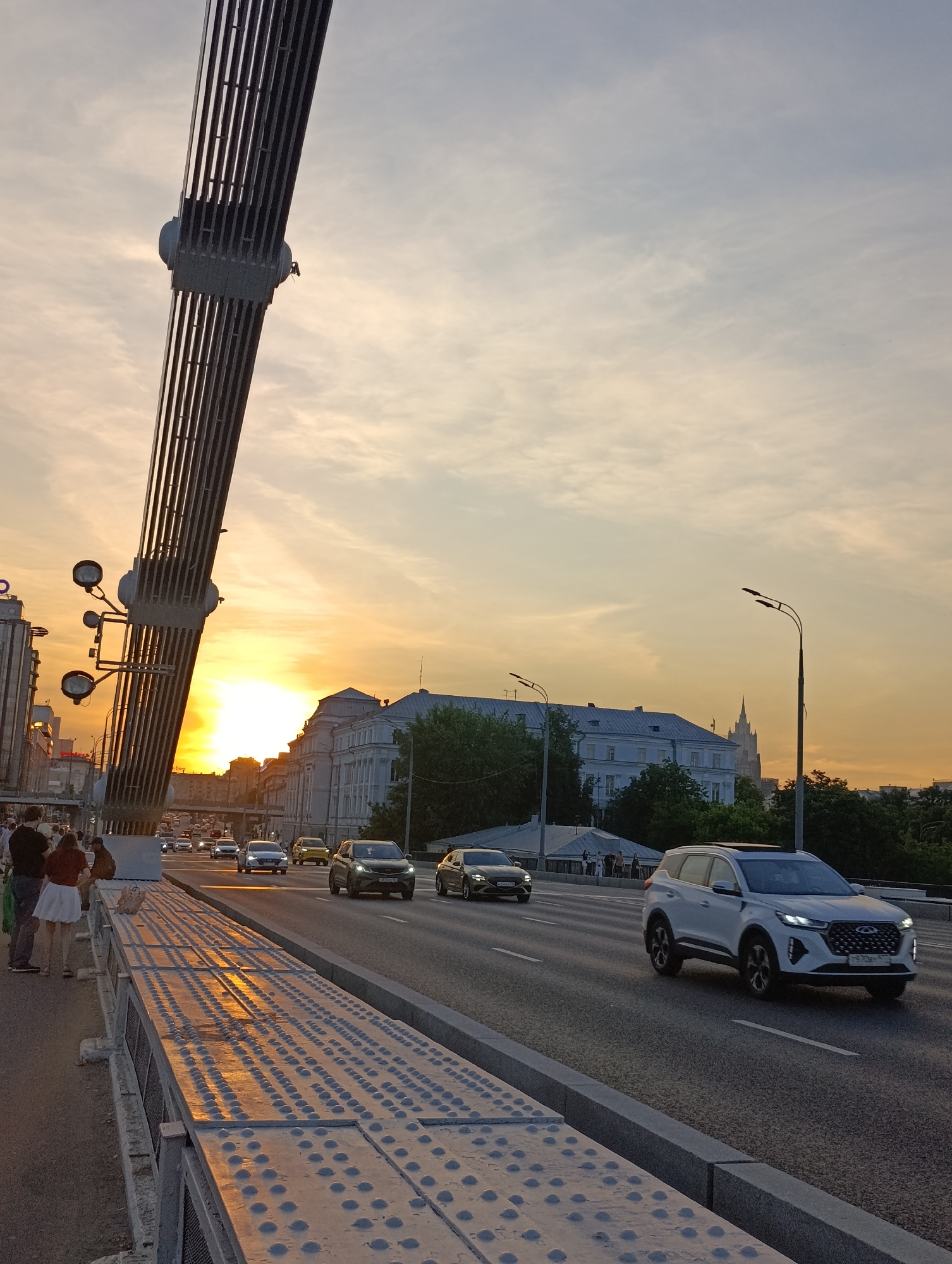This image captures an urban sunset scene on the Krymsky Bridge (Crimean Bridge) in Moscow, Russia. The perspective is from the pedestrian walkway on the bridge, looking west-southwest.
In the foreground on the left, a large, dark, ribbed metal structural element of the bridge, likely part of an arch or pylon, angles diagonally upwards and out of frame. Below this structure, the pedestrian walkway features light-colored metal panels with numerous raised circular studs, reflecting the warm glow of the setting sun. Approximately five to seven individuals are visible on this walkway; a man in a dark shirt and a woman in a white skirt and red top are prominent in the left mid-ground, facing away from the camera.
To the right of the walkway is a multi-lane asphalt roadway with vehicles moving away from the camera. The dominant vehicle in the right foreground is a white Chery Tiggo 8 Pro SUV. Further down the road, other vehicles include a dark SUV, a silver sedan, and a yellow car. Traffic appears moderate.
The sky is filled with high, wispy clouds, transitioning from a light blue at the top to a vibrant orange and yellow near the horizon, indicative of sunset. The sun itself is a bright, intense orange-yellow disc, partially obscured by distant buildings on the horizon in the left mid-ground.
On the far side of the river (beyond the roadway) and in the mid-distance, several light-colored, multi-story buildings with darker roofs are visible. Further in the distance, towards the right, the distinct silhouette of a Stalinist skyscraper with a prominent spire is visible, identified as the Main Building of the Ministry of Foreign Affairs of Russia. Two tall streetlights with modern fixtures line the right side of the roadway. Green foliage and trees are scattered across the distant landscape, particularly in front of and around the buildings.