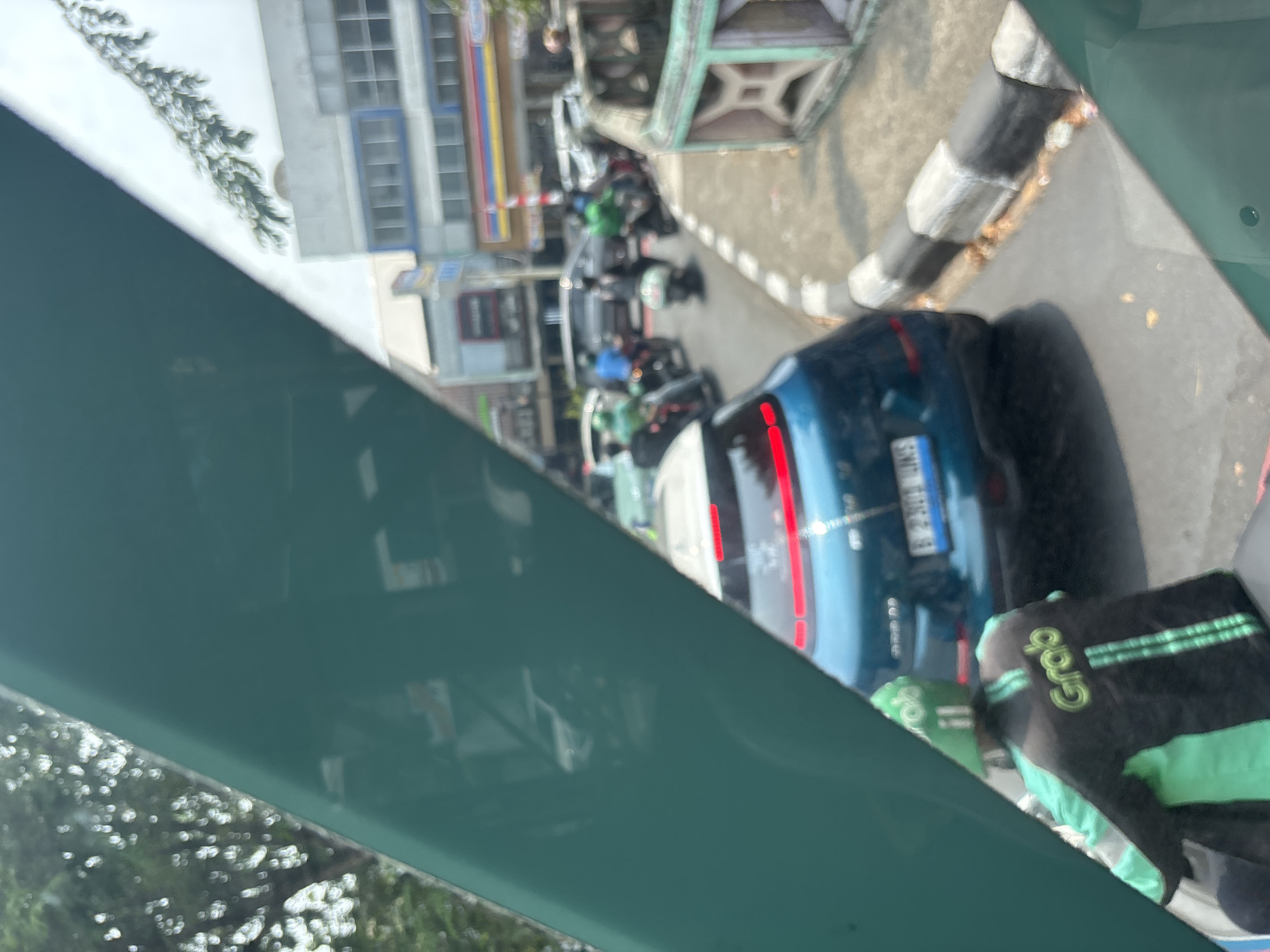 A first-person perspective shot, likely from a vehicle, captures a street scene in North Jakarta, Indonesia. In the foreground, a prominent teal-colored panel obscures the lower left portion of the frame. To the right, the rear of a teal-colored car with red taillights is visible. A person wearing a black jacket with green accents and the "Grab" logo on their back, along with a green helmet, is seated on a motorcycle in the lower left quadrant. Behind the teal car, a traffic jam is apparent, with several motorcycles and cars on the road. Many of the motorcycles have riders wearing green. The background reveals buildings lining the street, suggesting an urban environment. The weather appears to be daylight with some foliage visible.
