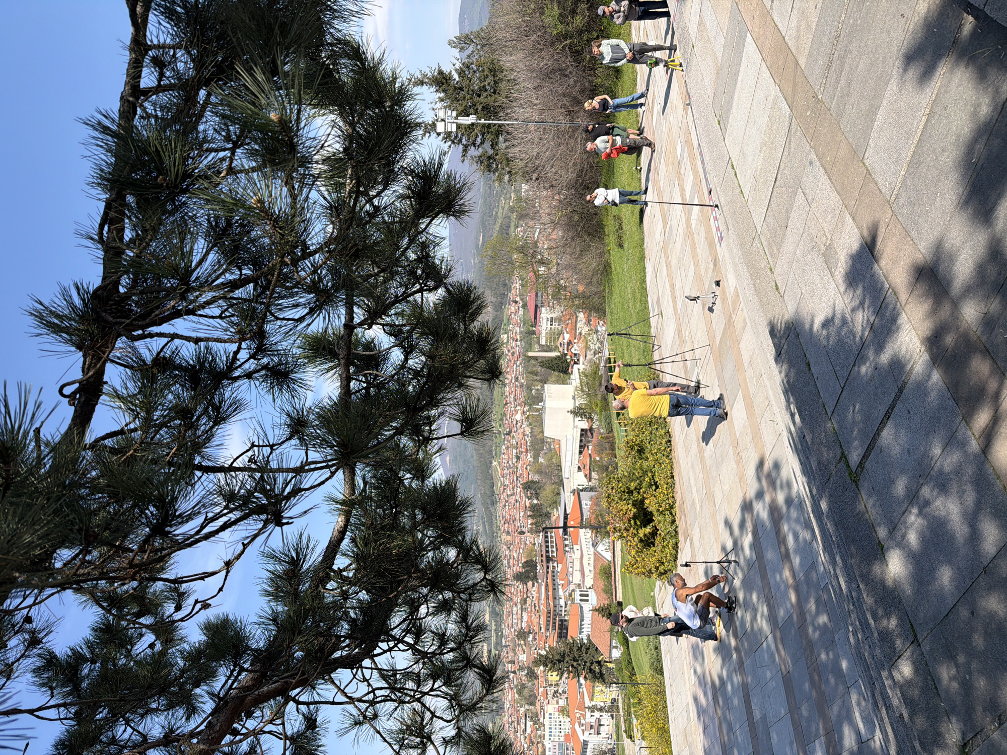The image depicts a group of people on a paved area overlooking the city of Panagyurishte, Bulgaria. The individuals are positioned along a stone walkway. Some appear to be engaged in some sort of work involving tall poles or tripods. A large pine tree is present on the left side of the frame, partially obscuring the view of the city. The city is visible in the background with buildings with orange roofs. The sky is blue with some clouds.