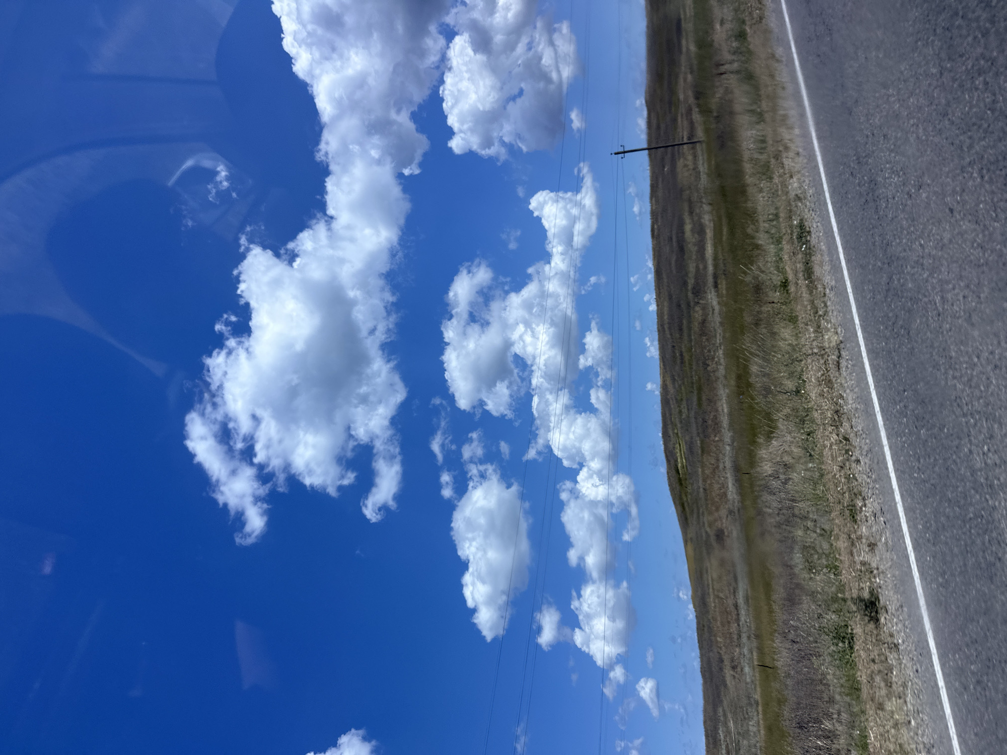 The image captures a roadside view of a grassy, rolling hill under a bright blue sky dotted with fluffy white clouds. Power lines are visible stretching across the horizon. In the foreground, a paved road with a white line marking its edge runs diagonally through the scene. The environment suggests a rural or semi-rural setting, possibly on the outskirts of Sennoy, Russia, given the dry vegetation and open landscape. There are no people or specific structures that would indicate a distinct urban location within Sennoy.