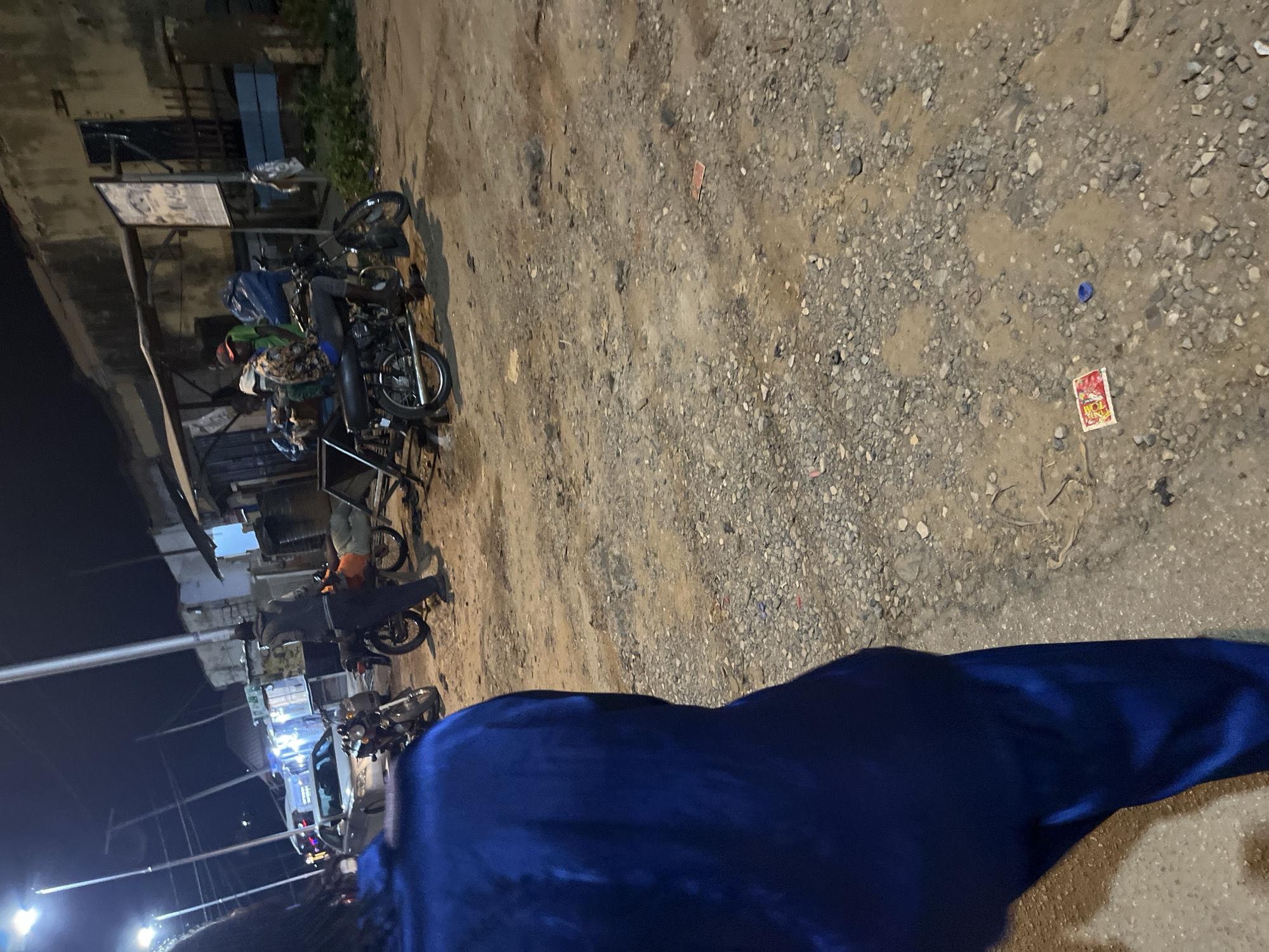 At night in a poorly lit area of Abuja, Nigeria, a person wearing a blue garment stands in the foreground, facing away from the camera. In the background, several motorcycles are parked on a dirt road. Some of the motorcycles are ridden by individuals, while others appear unoccupied. A rudimentary shelter with a corrugated metal roof is visible behind the motorcycles. The scene suggests a roadside gathering or a commercial area operating after dark.