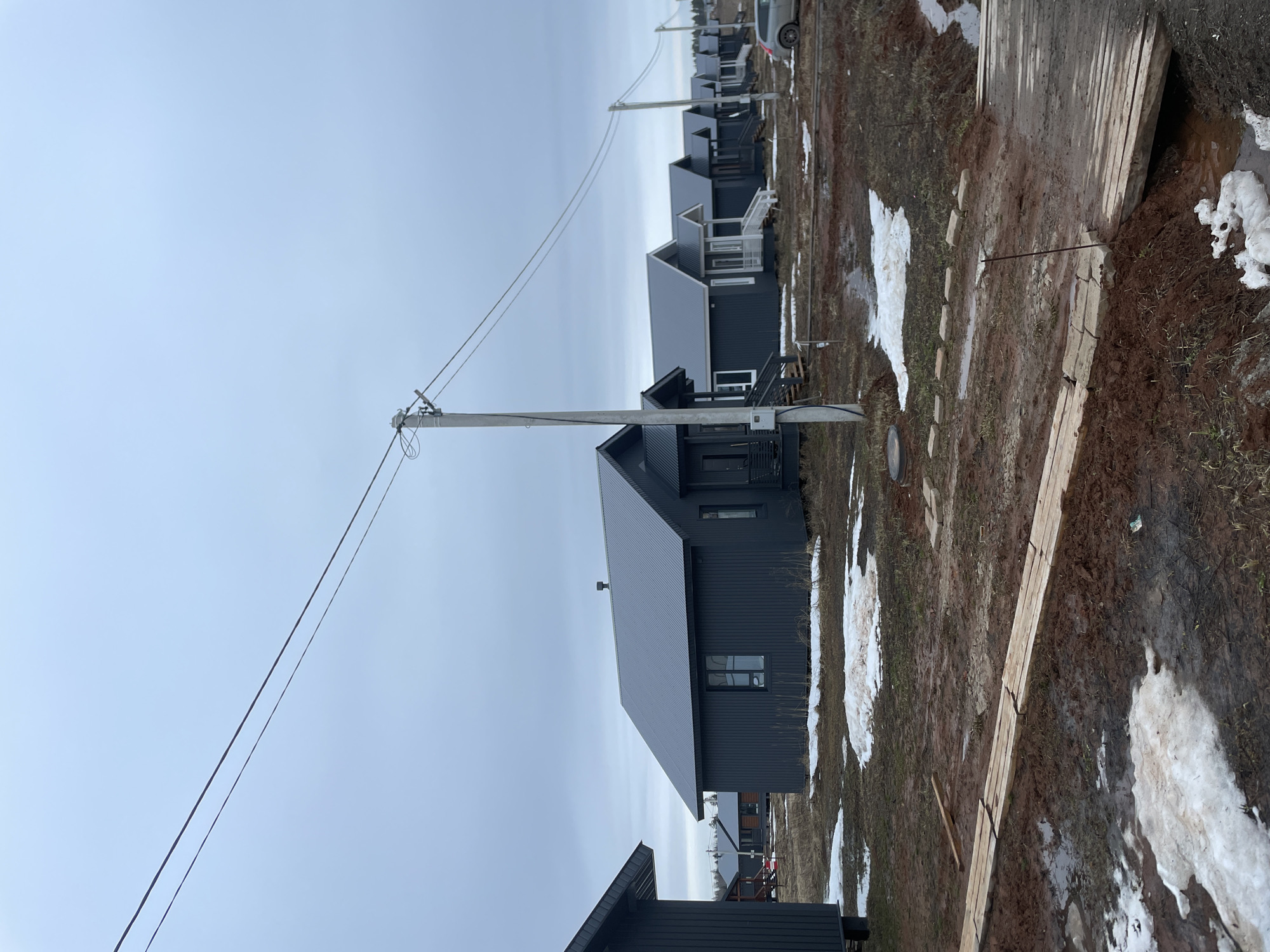 The image depicts a row of dark-colored houses, possibly newly constructed, in Подшивалово, Russia. The sky is overcast. In the foreground, the ground is muddy with patches of melting snow. Planks of wood are laid out on the ground. Utility poles with wires run along the road. A vehicle is visible in the background. No people are visible. The scene suggests a late winter or early spring day.