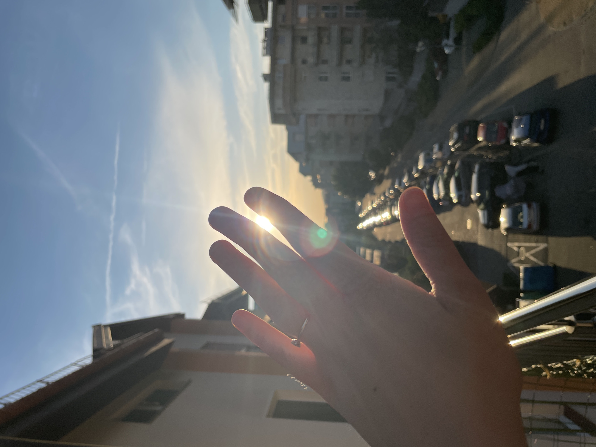 The media file displays a first-person perspective shot of a hand held up towards a bright sun, partially obscuring its rays. The hand has a ring on the ring finger. The scene is an urban environment with buildings and a street lined with parked cars visible in the background. The sky is blue with some wispy clouds, and the sun is low on the horizon, indicating it is either sunrise or sunset. Lens flare is prominent due to the direct sunlight. The overall mood appears peaceful and contemplative, with the sun's light creating a warm and serene atmosphere. There is no visible text in the image.