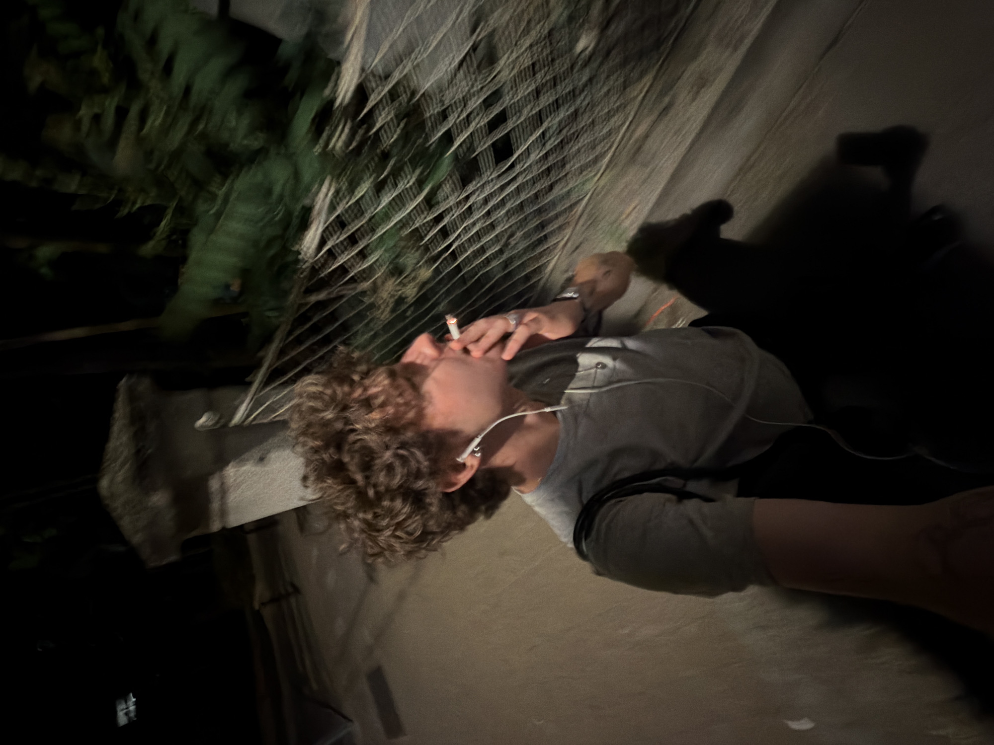 A person with curly hair, wearing headphones, is holding a cigarette near a wire fence. Green foliage is visible behind the fence. The scene appears to be outdoors, possibly at night, in a dark location with visible shadows. Vracar Urban Municipality, Serbia cannot be confirmed as the location.