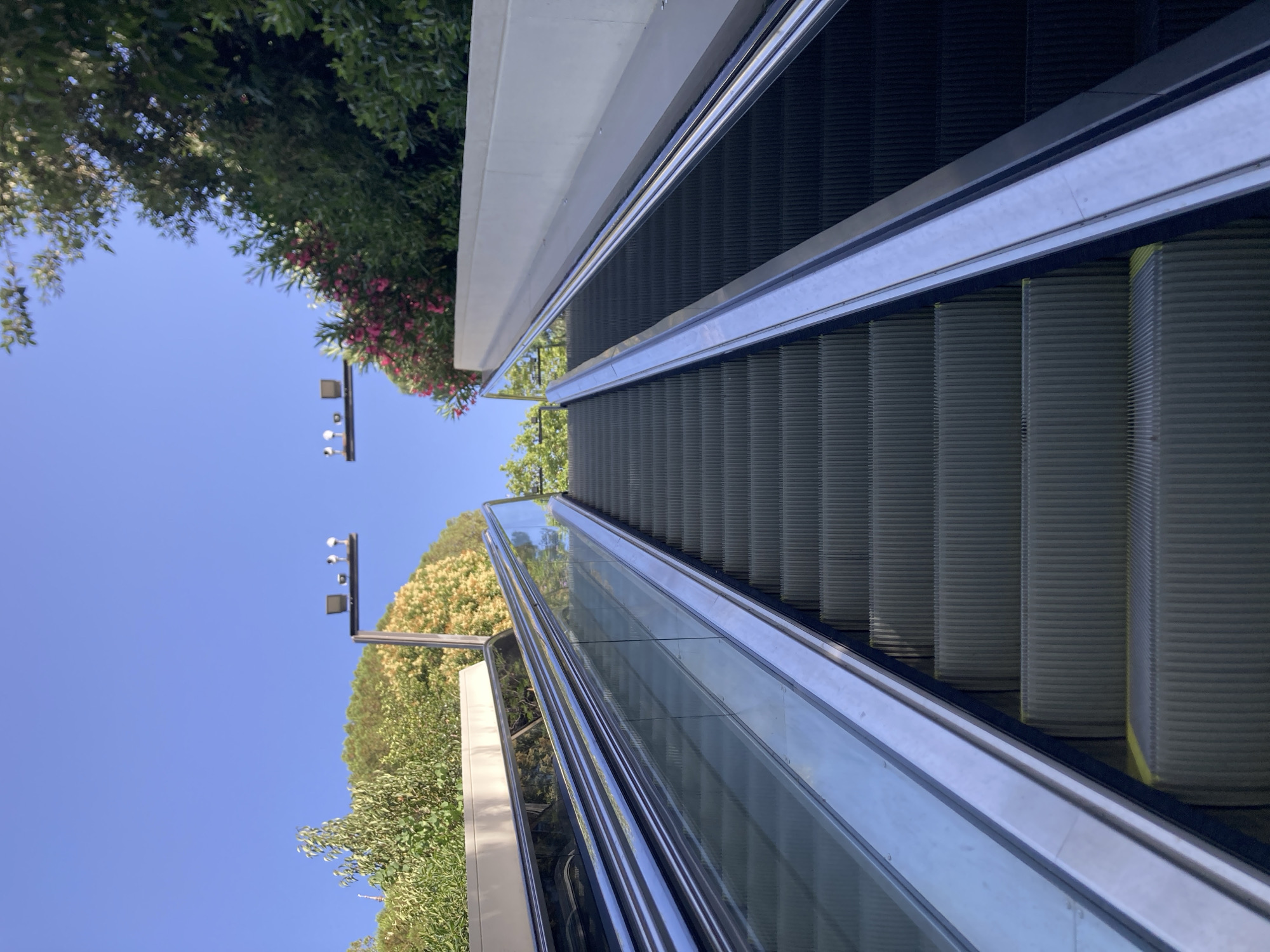 An outdoor escalator in Barcelona, Spain, ascends towards a clear blue sky. The escalator is flanked by glass and metal railings. Lush green trees and flowering bushes line the sides of the escalator, with a hint of yellow foliage visible on the left. Two lampposts with multiple light fixtures are positioned above the escalator, one on each side, amidst the greenery. The escalator steps are dark grey, and the surrounding environment is well-lit by natural sunlight.