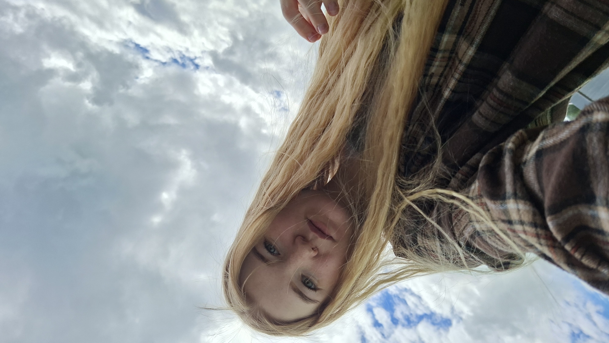 A low-angle portrait captures a young person with long, flowing blonde hair looking down towards the camera. The subject's face, with light-colored eyes and a neutral expression, is visible in the lower half of the frame. A hand is present at the very top of the image. The person is wearing a brown, beige, and red plaid flannel shirt, seen on the right side and hinted at on the left.
The entire background is a cloudy sky, featuring shades of white and grey with patches of blue visible through the clouds, especially towards the upper and lower right. The perspective suggests the person is positioned above the viewer or looking down from a height. The lighting indicates it is daytime with an overcast sky. No text is visible in the image. This scene is set in Большой Букор, Russia.