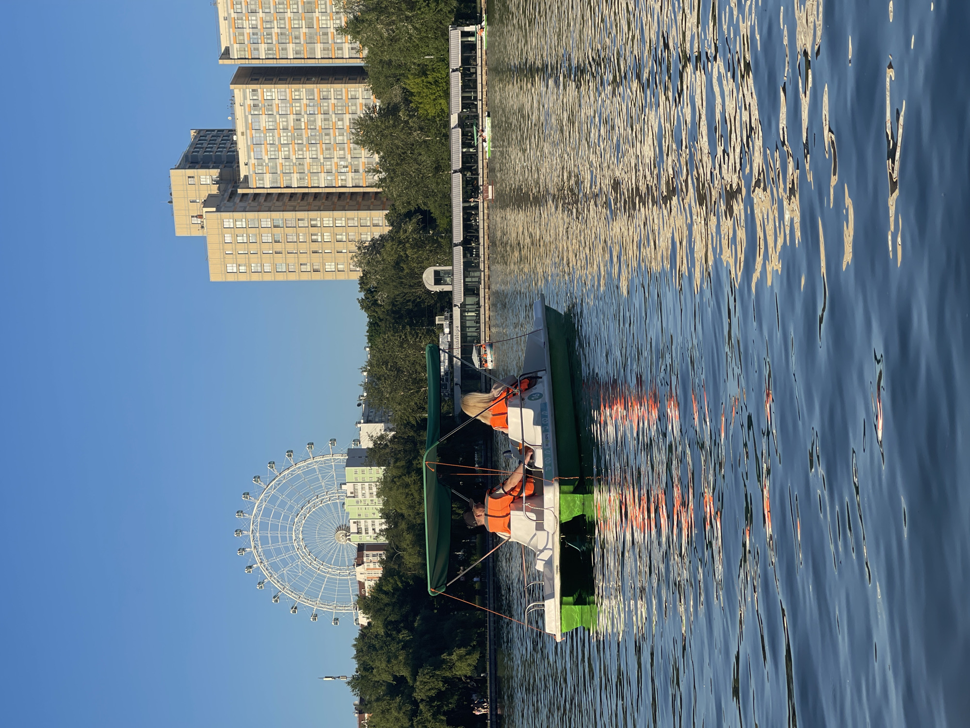 The location is an urban park with a large pond or lake in an unidentified city, likely within a Russian-speaking country given the text "Акватория" visible on a building. The scene depicts a bright, clear summer late afternoon or early evening.
In the foreground, a green and white pedal boat with a green canopy moves diagonally away from the viewer on the rippled water. Two individuals occupy the boat, both wearing orange life vests. The person on the left is a male wearing a dark cap and a dark shirt, positioned as if steering. The person on the right is a female with blonde hair, wearing a light-colored top, also facing forward.
The midground features a calm body of water reflecting the sky and distant architecture. On the distant left shore, a long, low-rise building with large windows and dark awnings is visible, partially labeled with the Russian text "Акватория" (Aquatoria). Several individuals are present on the shore and near this building. A line of lush green trees extends along the shoreline. Other smaller pedal boats are sparsely scattered across the water in the distance.
In the background, against a clear blue sky, a prominent white Ferris wheel with multiple gondolas is visible on the left side, rising above the tree line. To the right, a cluster of tall, modern residential buildings with light-colored, multi-story facades and numerous windows dominates the skyline, their structural reflections shimmering on the water's surface. The golden sunlight indicates a late afternoon or early evening setting.