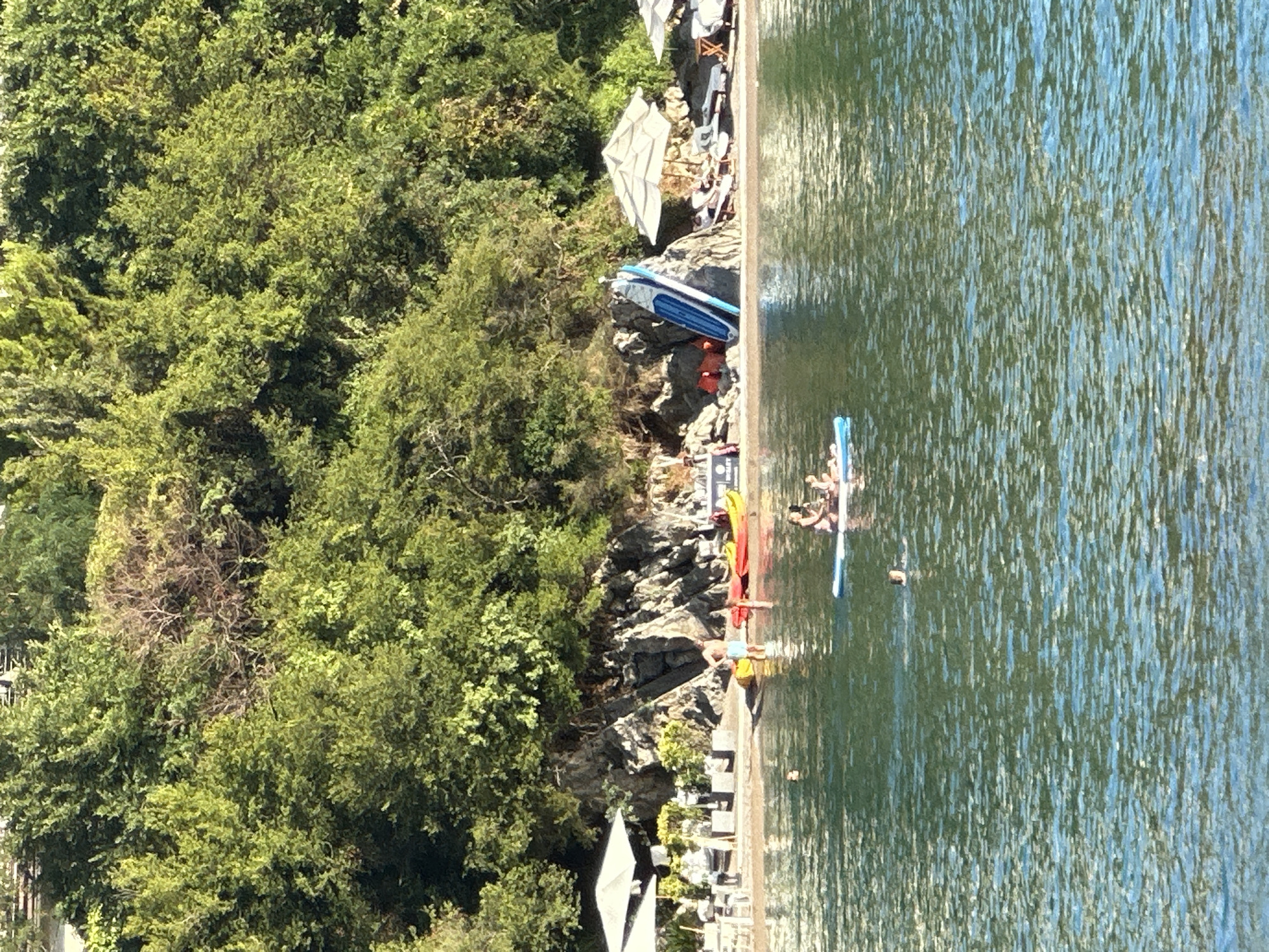 The media file depicts a sun-drenched scene at a waterfront, likely in Budva, Montenegro, given the context. A lush, green hillside teeming with trees forms the backdrop on the left side of the frame. The foreground is dominated by the tranquil, rippling blue water of the sea or a large lake.
Several objects are scattered along the shoreline and in the water. On the rocky shore, a blue and white kayak or paddleboard rests, accompanied by orange life vests and other watercraft. Further along the shore, a row of red kayaks and a yellow kayak are visible, suggesting a rental or recreational area.
In the water, a person is seen on a paddleboard, seemingly enjoying a leisurely activity. To the right, another paddleboard is occupied by a group of individuals, their forms blurred by the distance and water's reflection. The scene suggests a warm, sunny day, likely during daylight hours, with clear weather conducive to outdoor water activities. The overall mood is relaxed and recreational, with a focus on enjoying the natural beauty of the water and the surrounding greenery. There are no visible text elements in the media file.