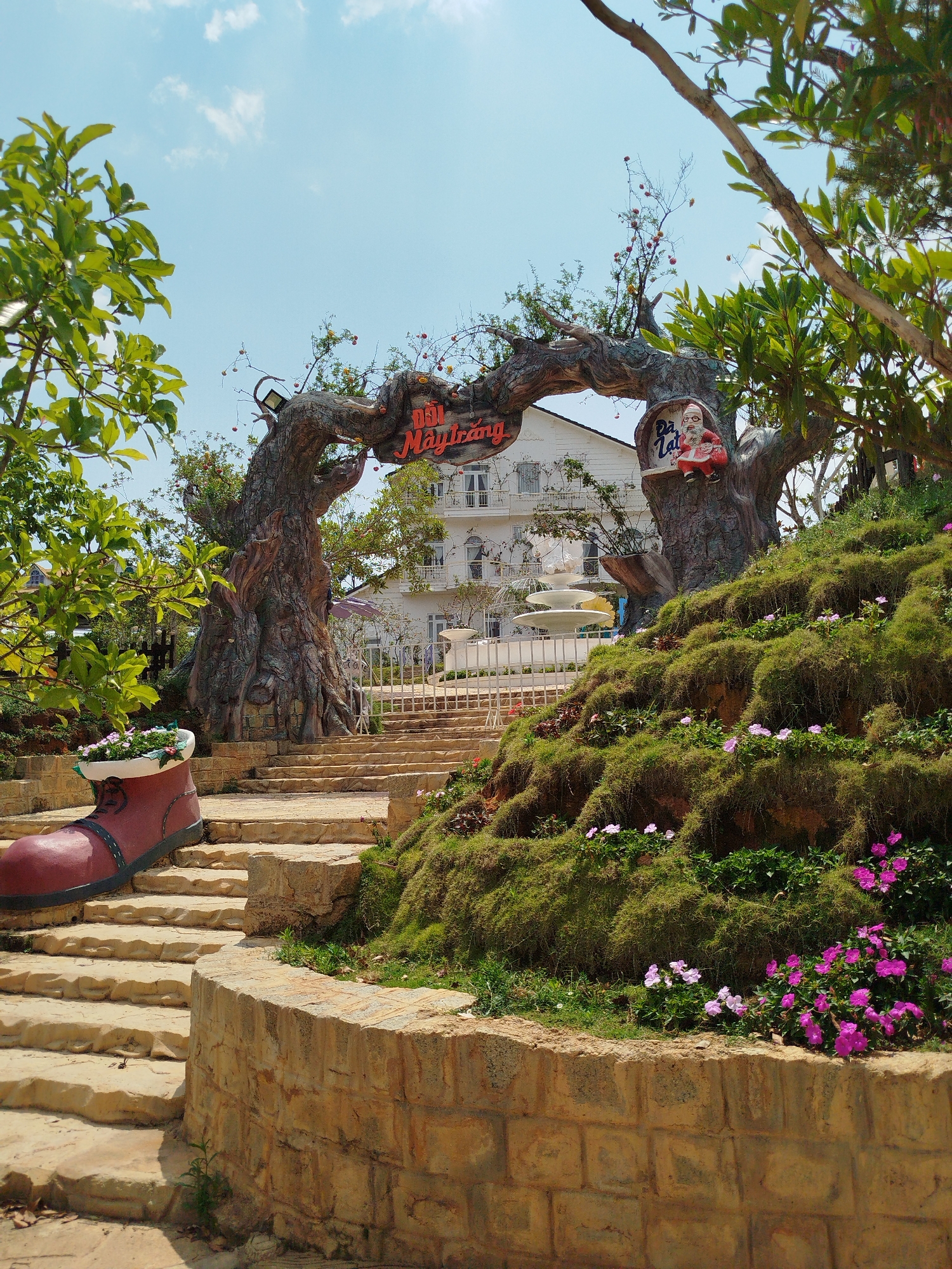 The image captures a garden scene in Lang Biang - Da Lat, Vietnam. A stone staircase leads up to a white building with multiple windows and a fountain. An elaborate tree-trunk archway with signage marks the entrance. A large, red, boot-shaped planter sits on the left side of the steps. The landscape features terraced greenery with pink and white flowers. A Santa Claus figure is perched on one of the tree trunks. The sky is bright blue with scattered clouds.